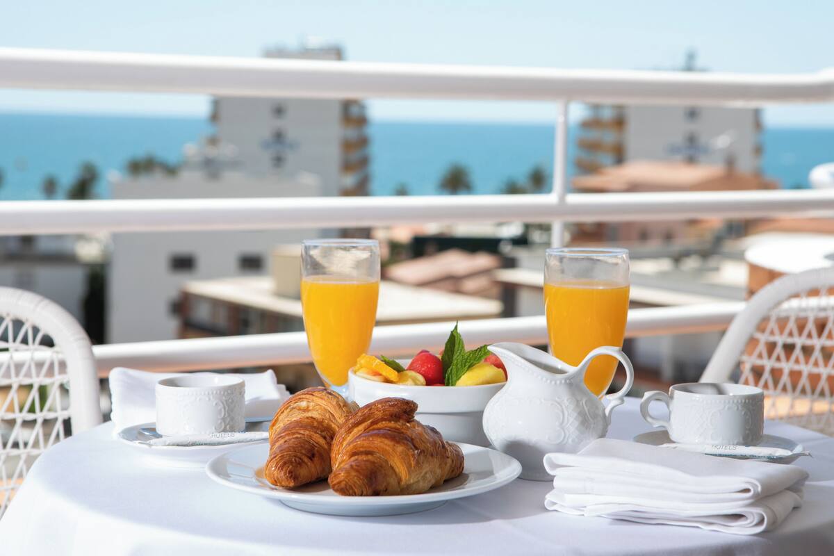 A breakfast table with croissants, fruit, and orange juice on a balcony overlooking the sea.