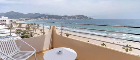 Un balcon avec une table et des chaises blanches, donnant sur une plage et des montagnes.