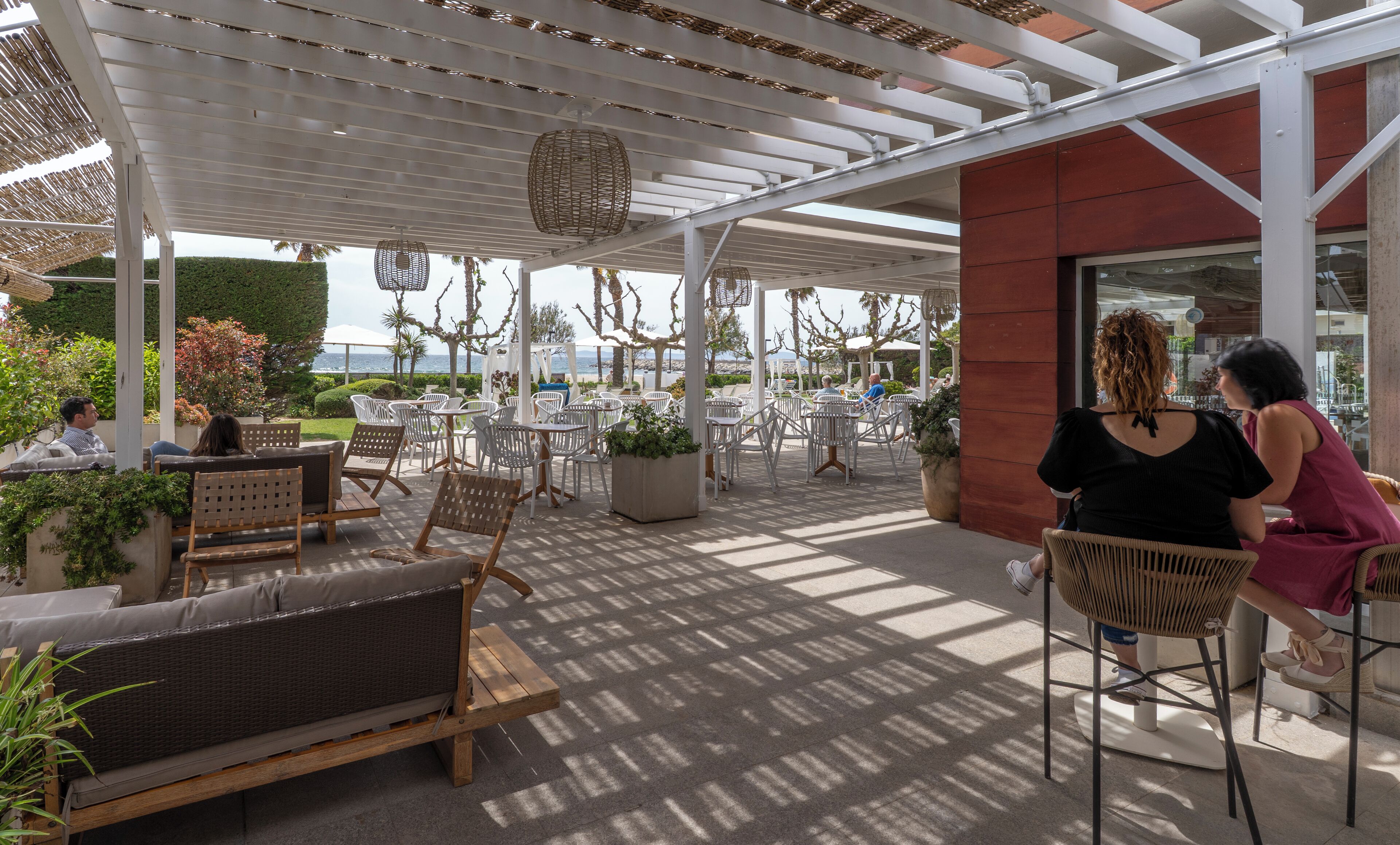 An outdoor patio area with seating arrangements, including sofas and chairs, under a covered structure with a view of the sea.