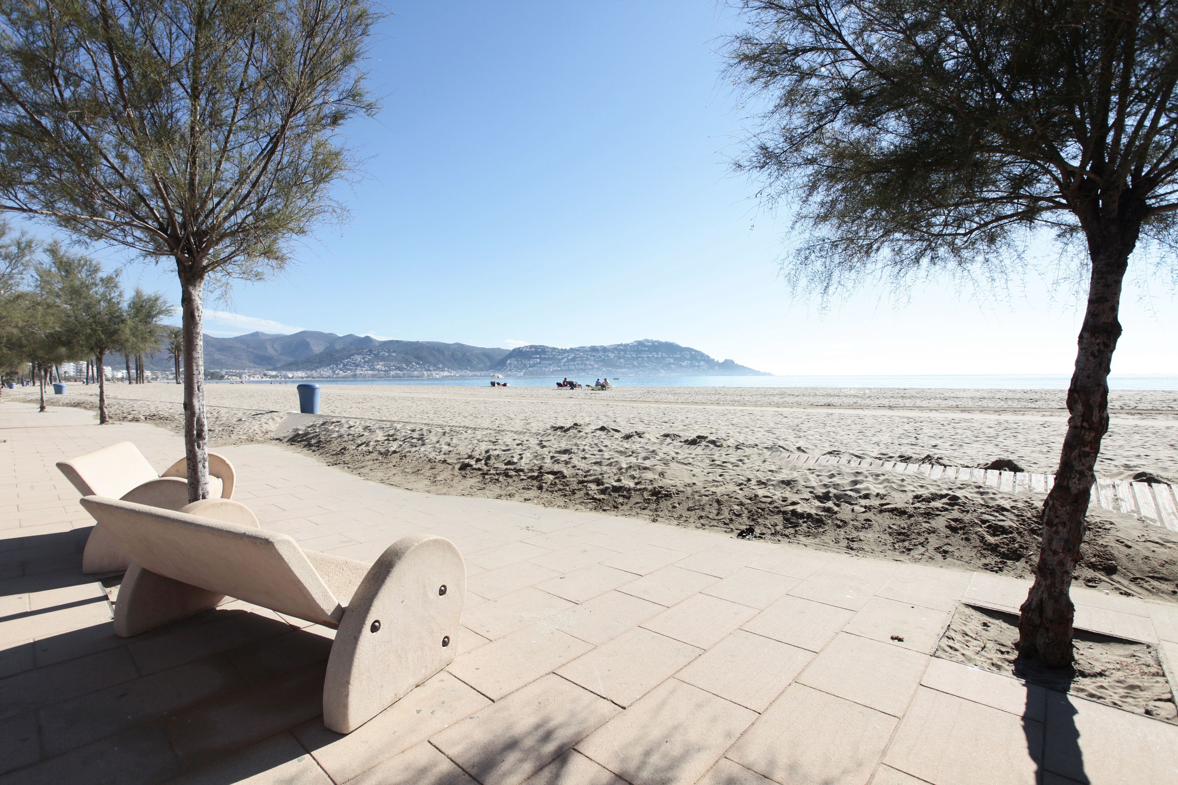 Uma passarela pavimentada com bancos e árvores, que leva a uma praia com montanhas ao longe.