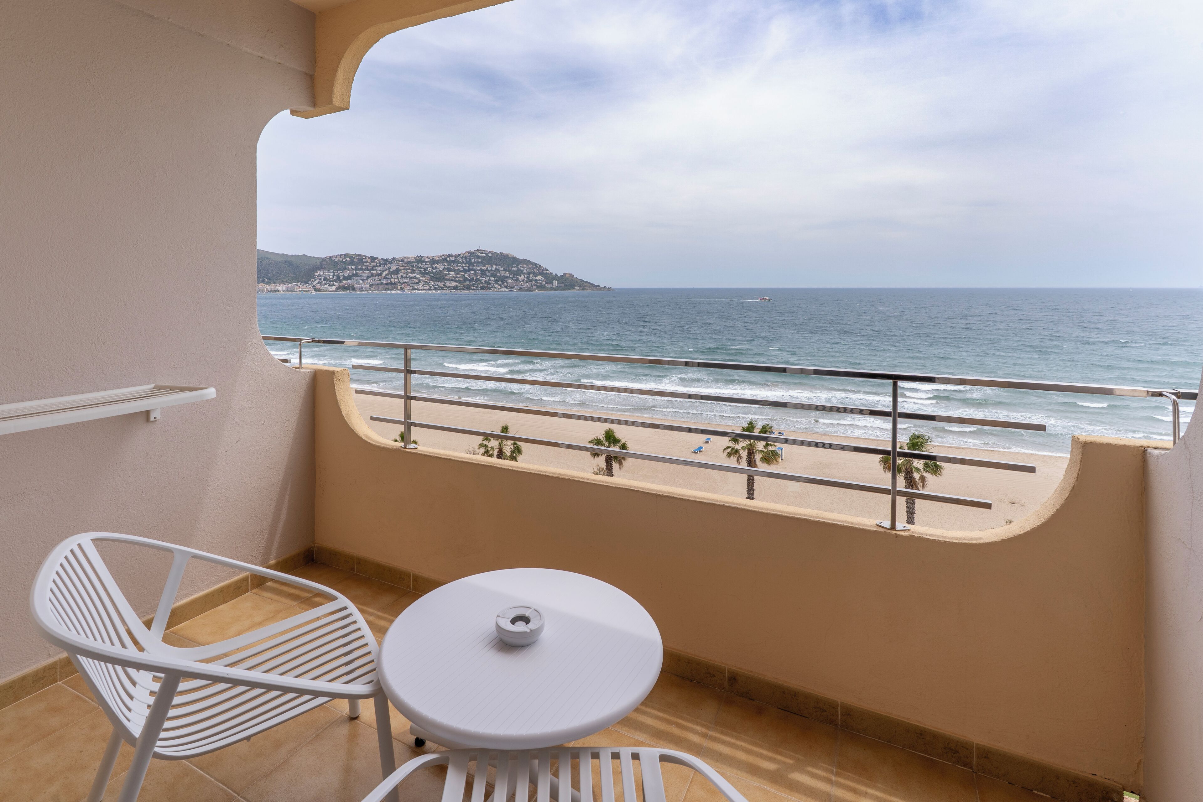 A balcony with a white table and chair, overlooking a beach with palm trees and a distant mountain.