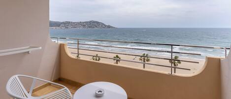 A balcony with a white table and chair, overlooking a beach with palm trees and a distant mountain.