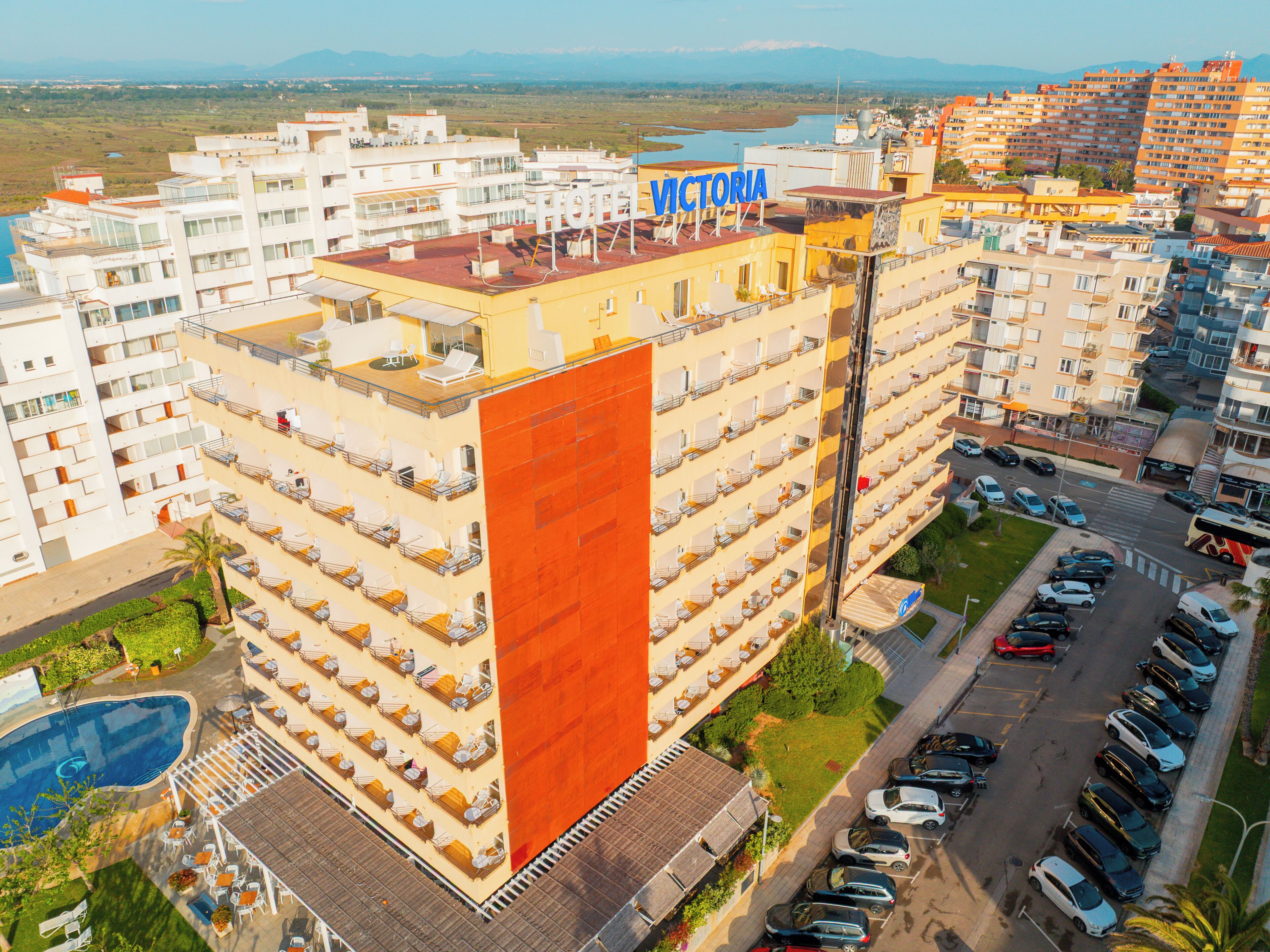 A multi-story hotel with a rooftop pool, a sign reading 'VICTORIA HOTEL,' and a parking lot with several cars.