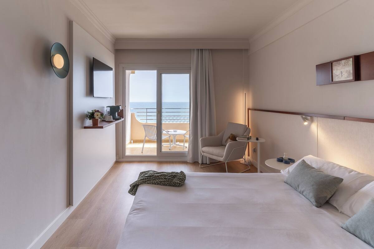 Une chambre d’hôtel avec un lit, une chaise, une petite table, un téléviseur et une vue sur la mer, accessible par une porte coulissante.