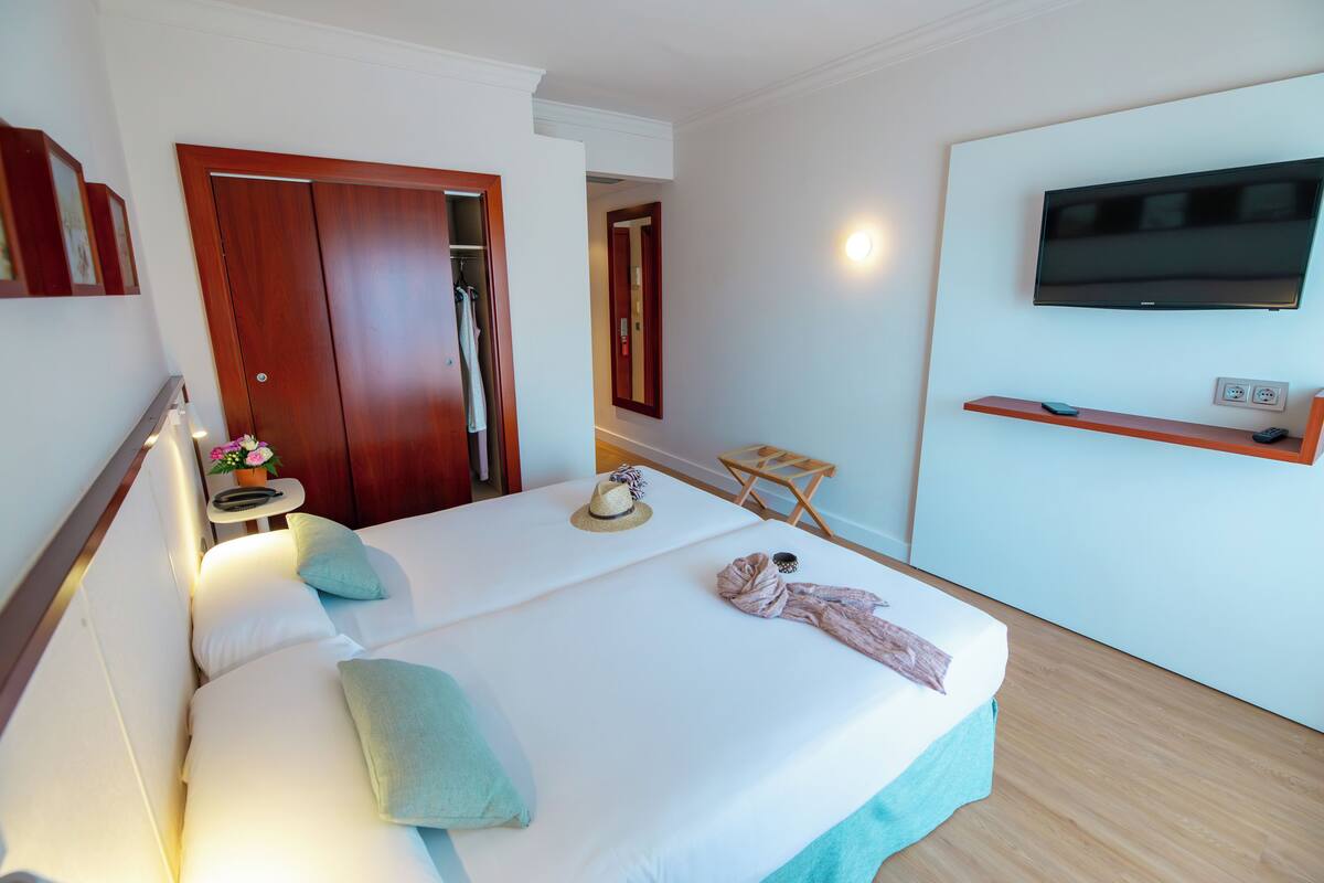 Une chambre d’hôtel avec un lit, une télévision, une armoire en bois et une petite table sur laquelle est posé un vase de fleurs.