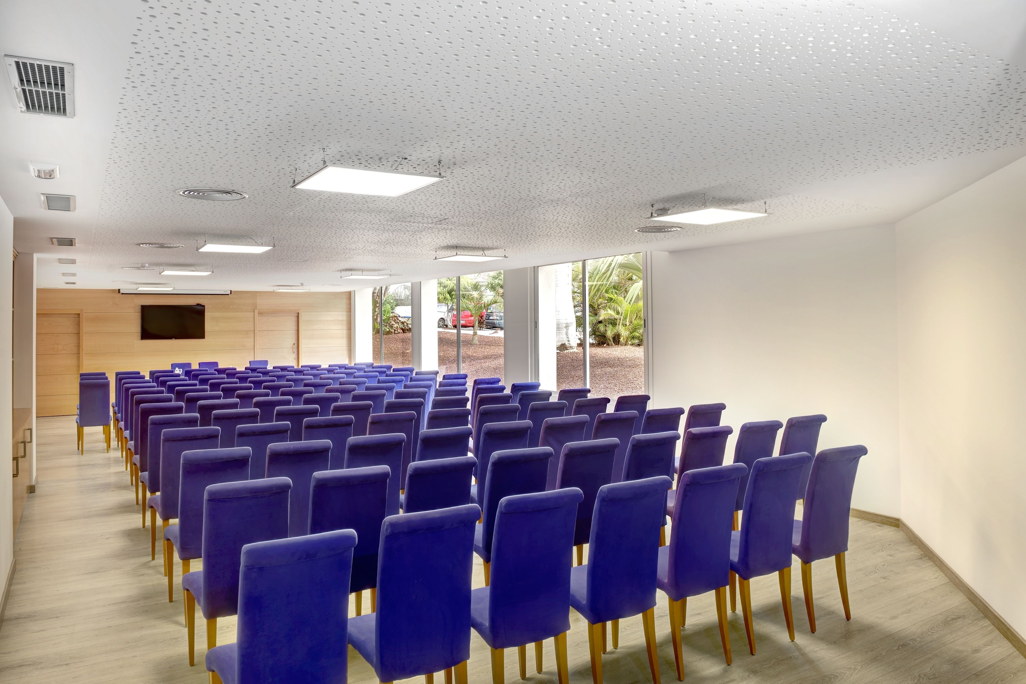 Un salón de conferencias con hileras de sillas moradas, un panel de pared de madera y ventanas amplias que dan a vistas de palmeras.