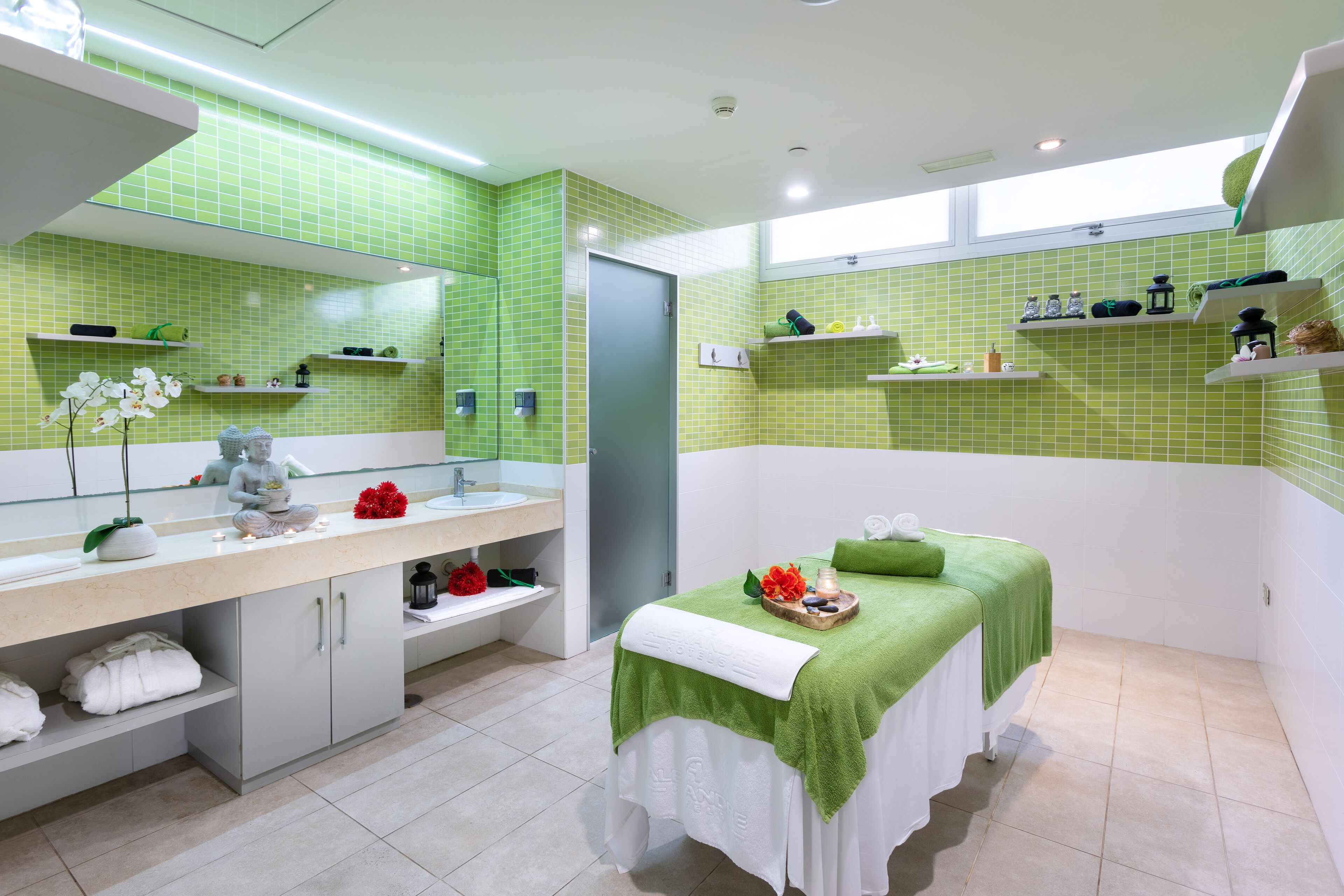 Un moderno cuarto de spa con una paleta de colores verde y blanco, un espejo grande, una camilla de masaje y estantes con artículos de aseo.