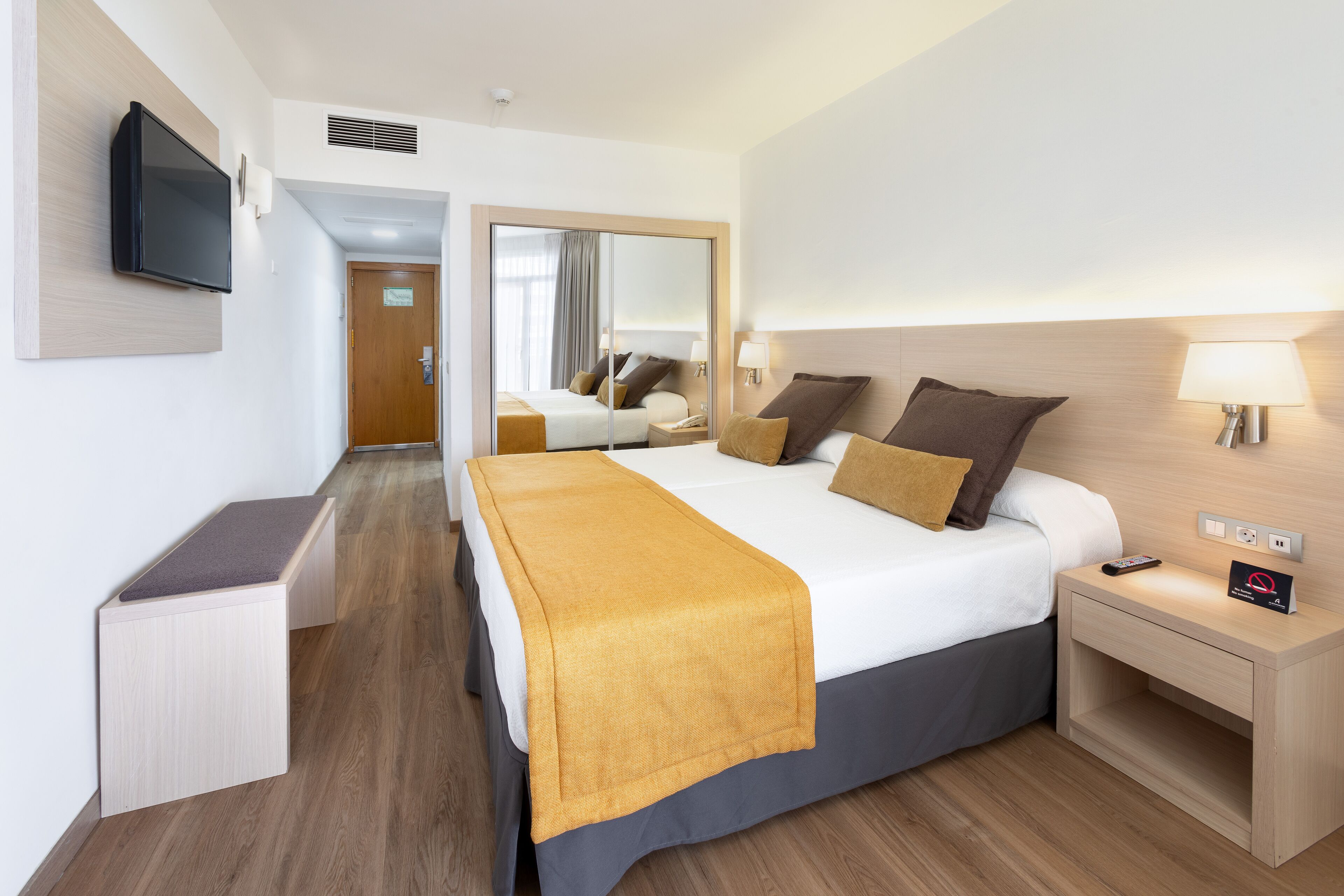 Una habitación de hotel con cama, mesita de noche, lámpara y televisor montado en la pared.