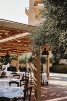 Un área de comedor al aire libre con mesas cubiertas con manteles blancos, estructuras de pérgola de madera y olivos.