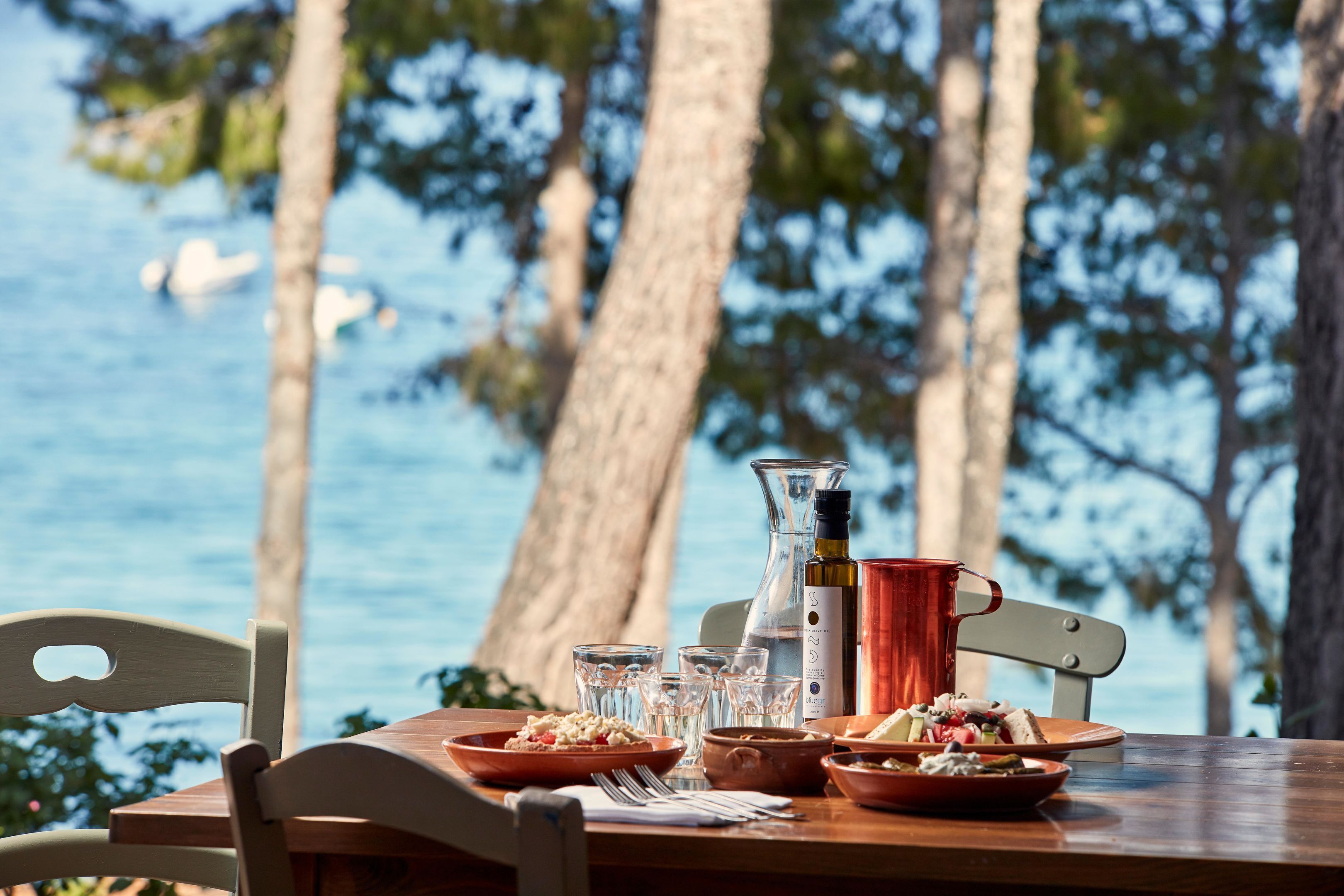 A table set with plates of food, glasses, and a bottle, overlooking a body of water with trees in the background.