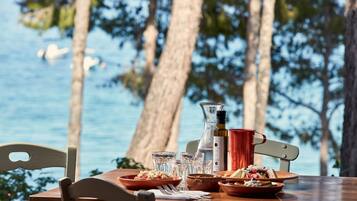 A table set with plates of food, glasses, and a bottle, overlooking a body of water with trees in the background.