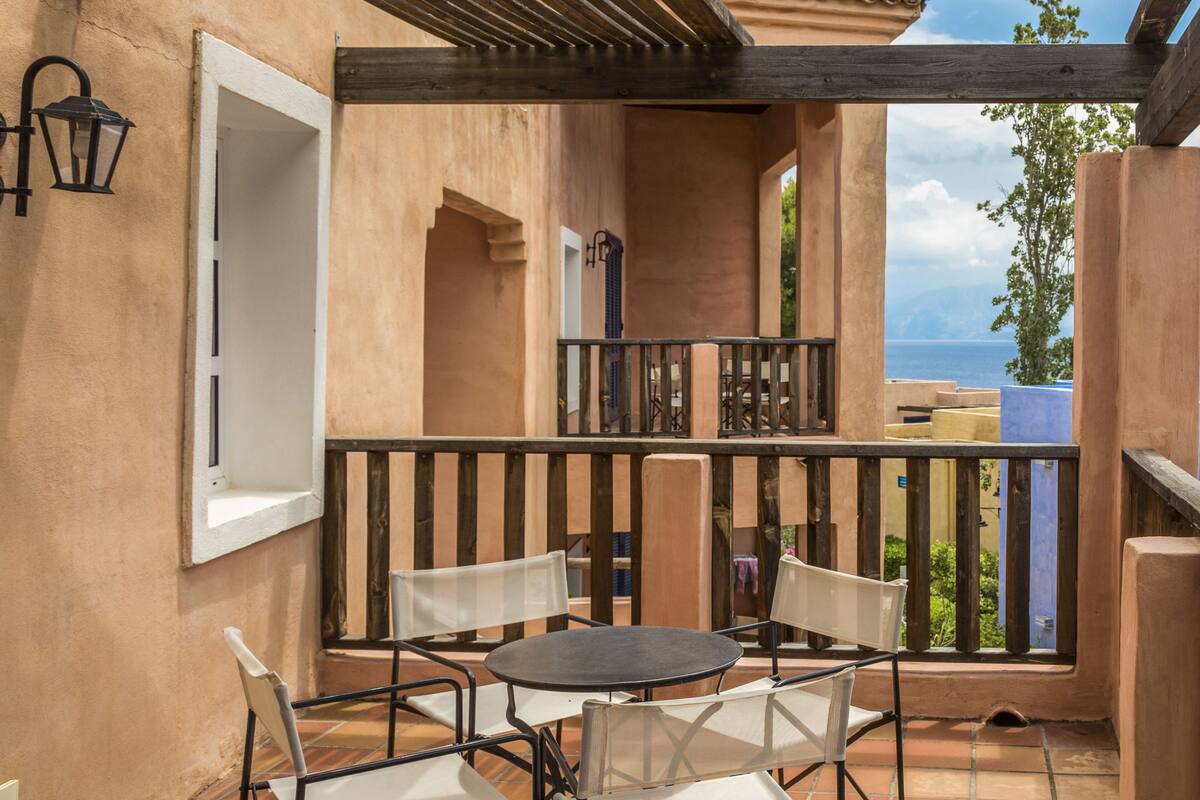 A terrace with a round table and chairs, a wooden railing, and a view of the sea.