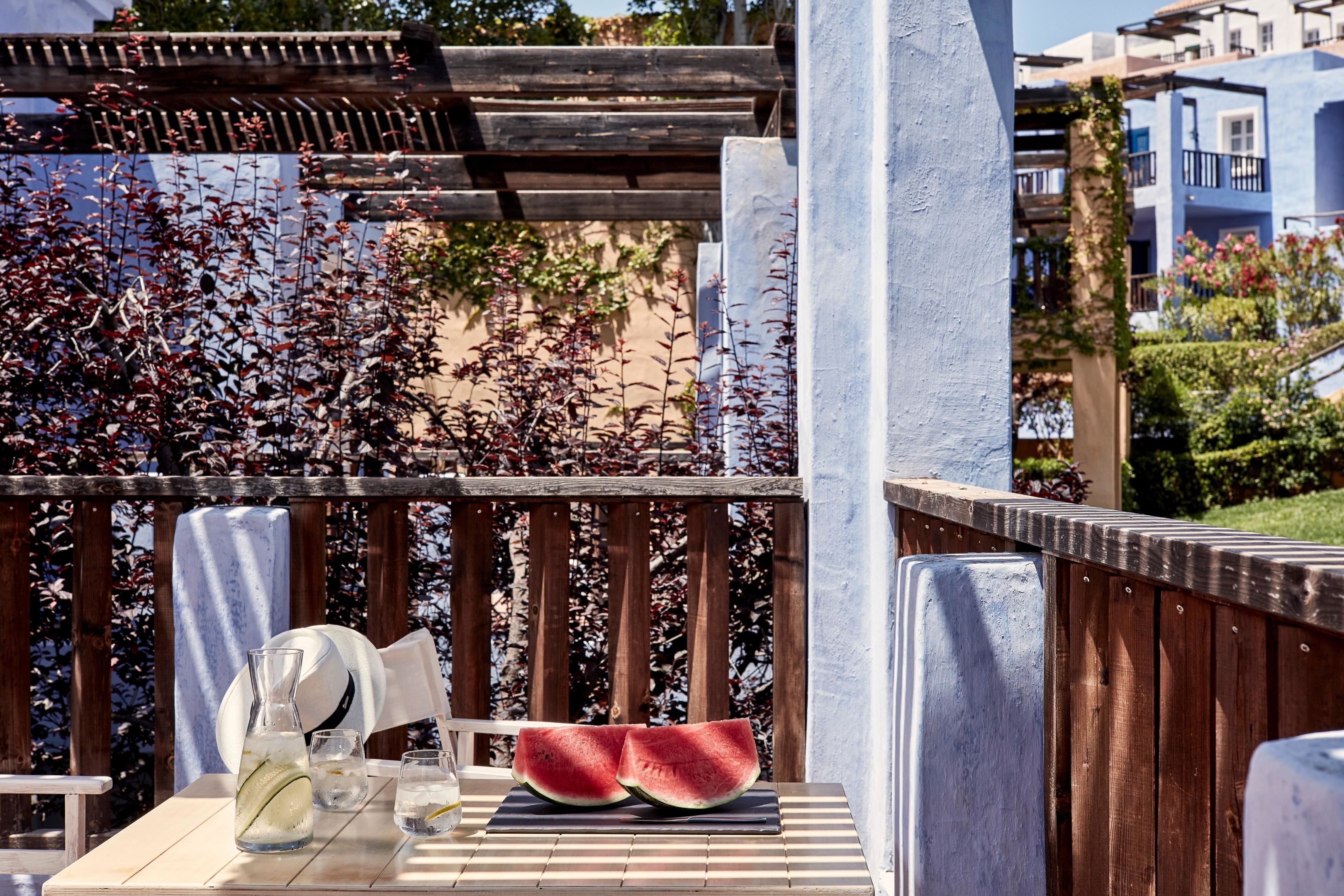 Une terrasse en bois avec une table dressée pour deux personnes, agrémentée d’une pastèque et d’une carafe de limonade.