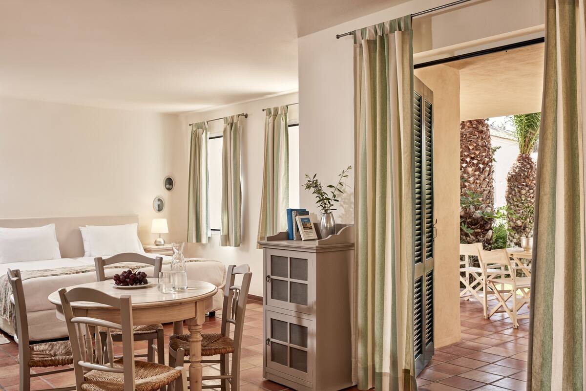 A hotel room with a bed, a dining table with chairs, a cabinet, and a view of the outdoor area.