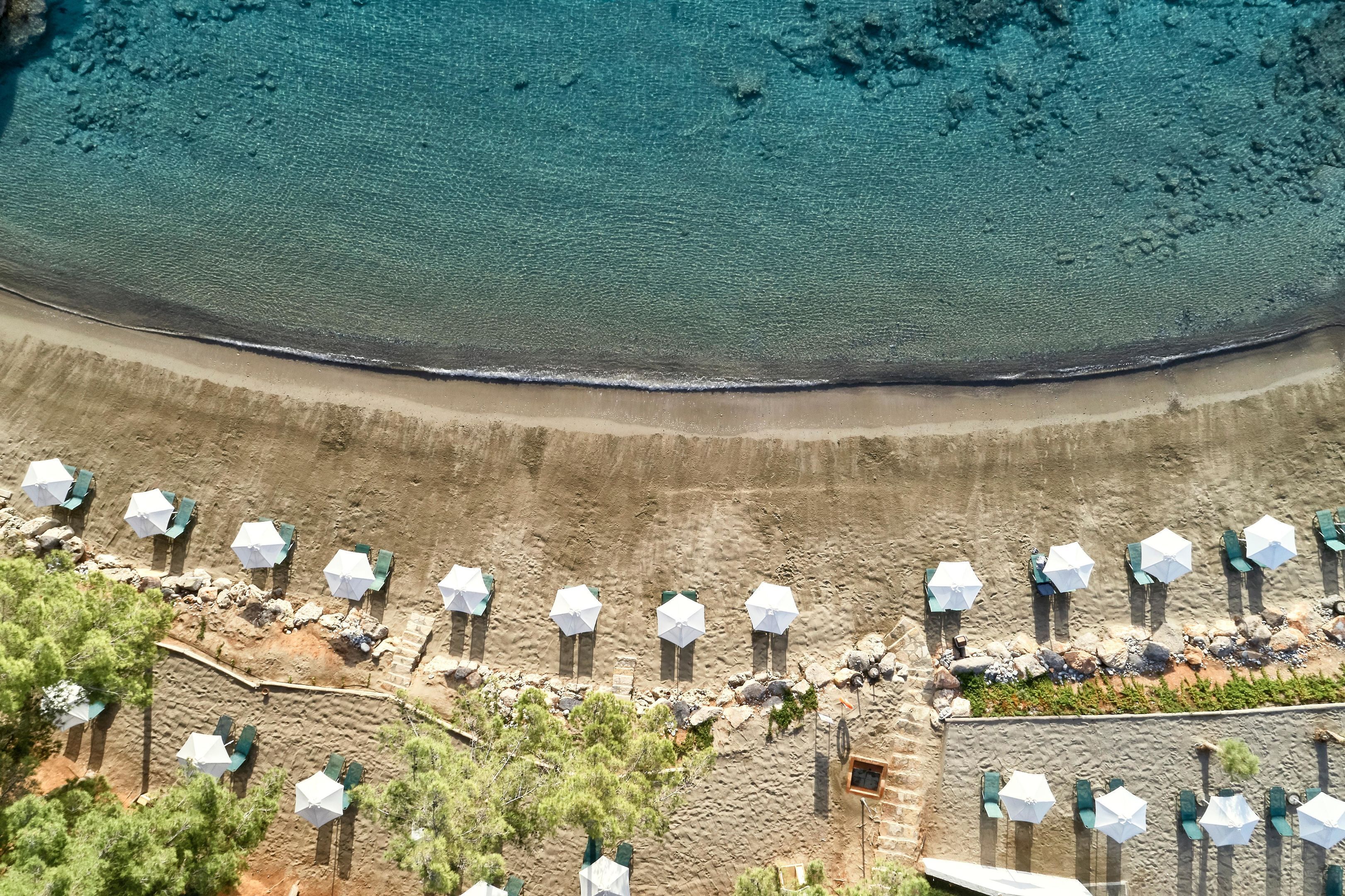 Une vue aérienne d’une plage bordée de rangées de parasols et de chaises, avec une vue dégagée sur l’océan et un rivage rocheux.