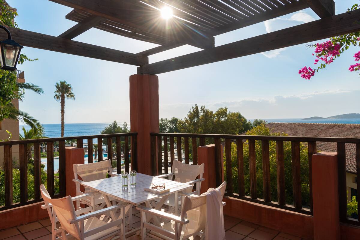 A wooden deck with a table and chairs, overlooking a coastal view with palm trees and a clear sky.