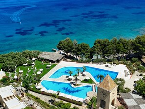 Ovenudblik over et resort med en stor swimmingpool, et klokketårn og et strandområde.