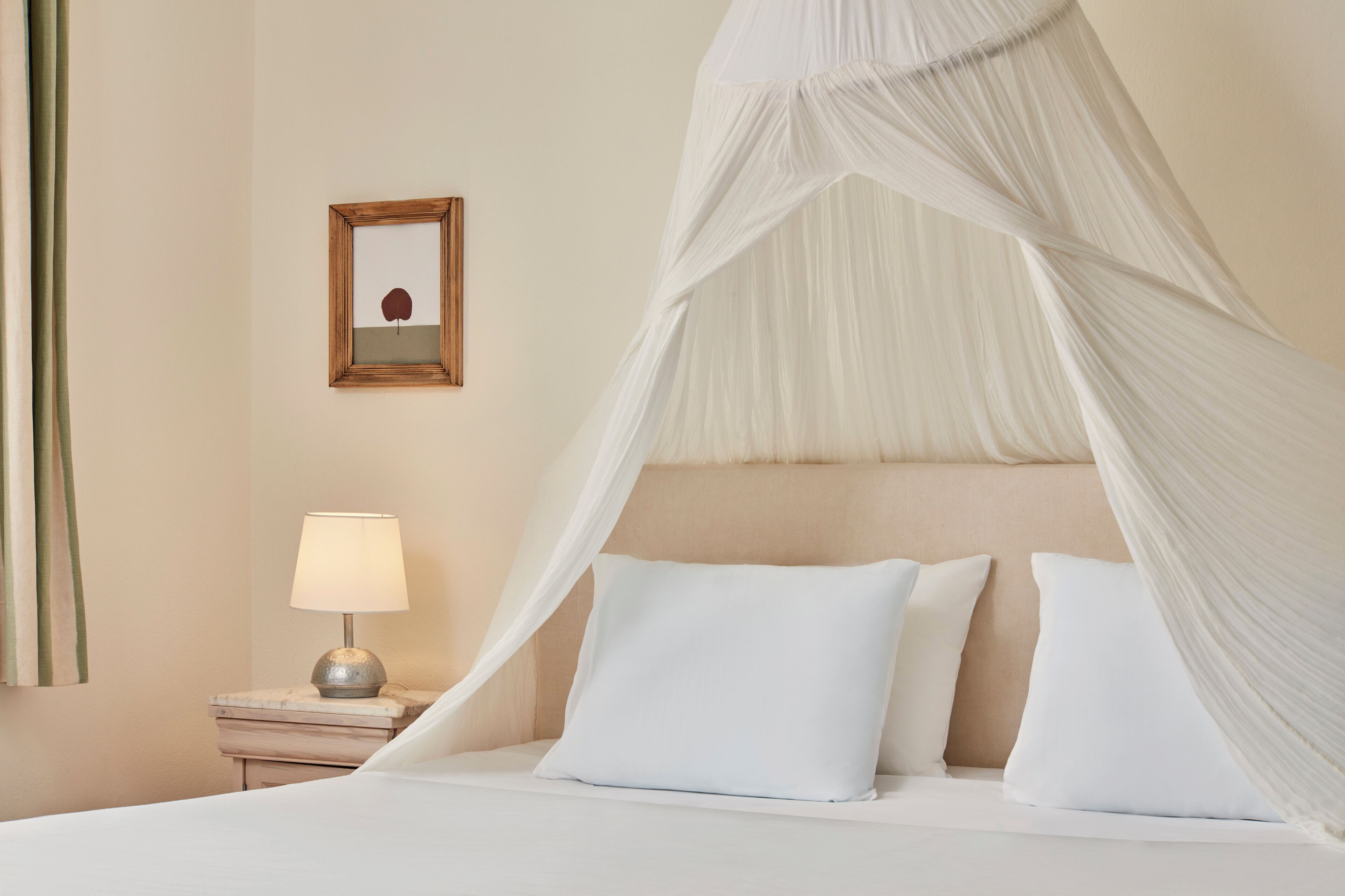 Una cama con ropa blanca y dosel, una mesita de noche con lámpara, y un cuadro en la pared.