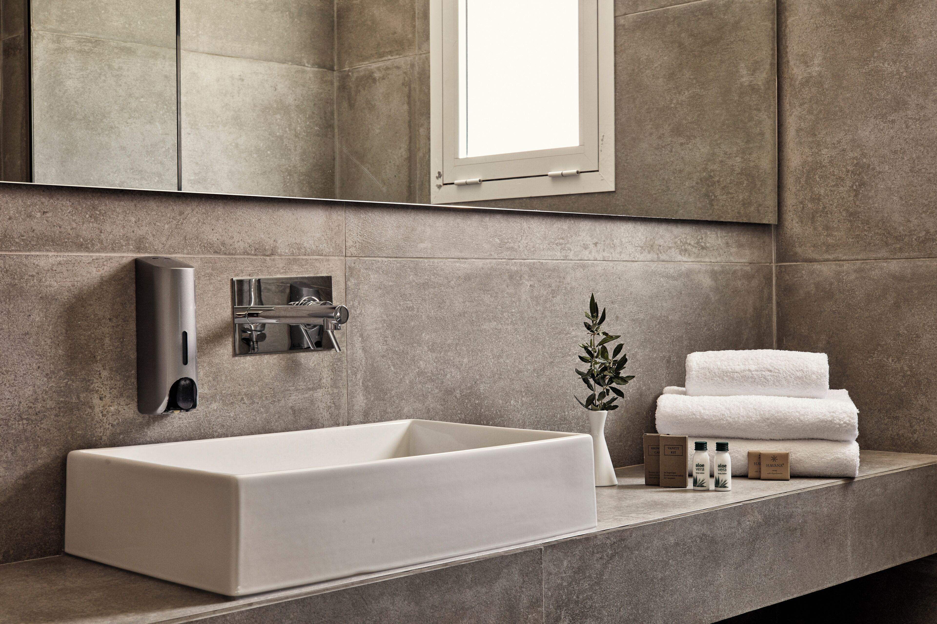 A modern bathroom with a rectangular sink, a wall-mounted faucet, a small potted plant, and neatly stacked towels.