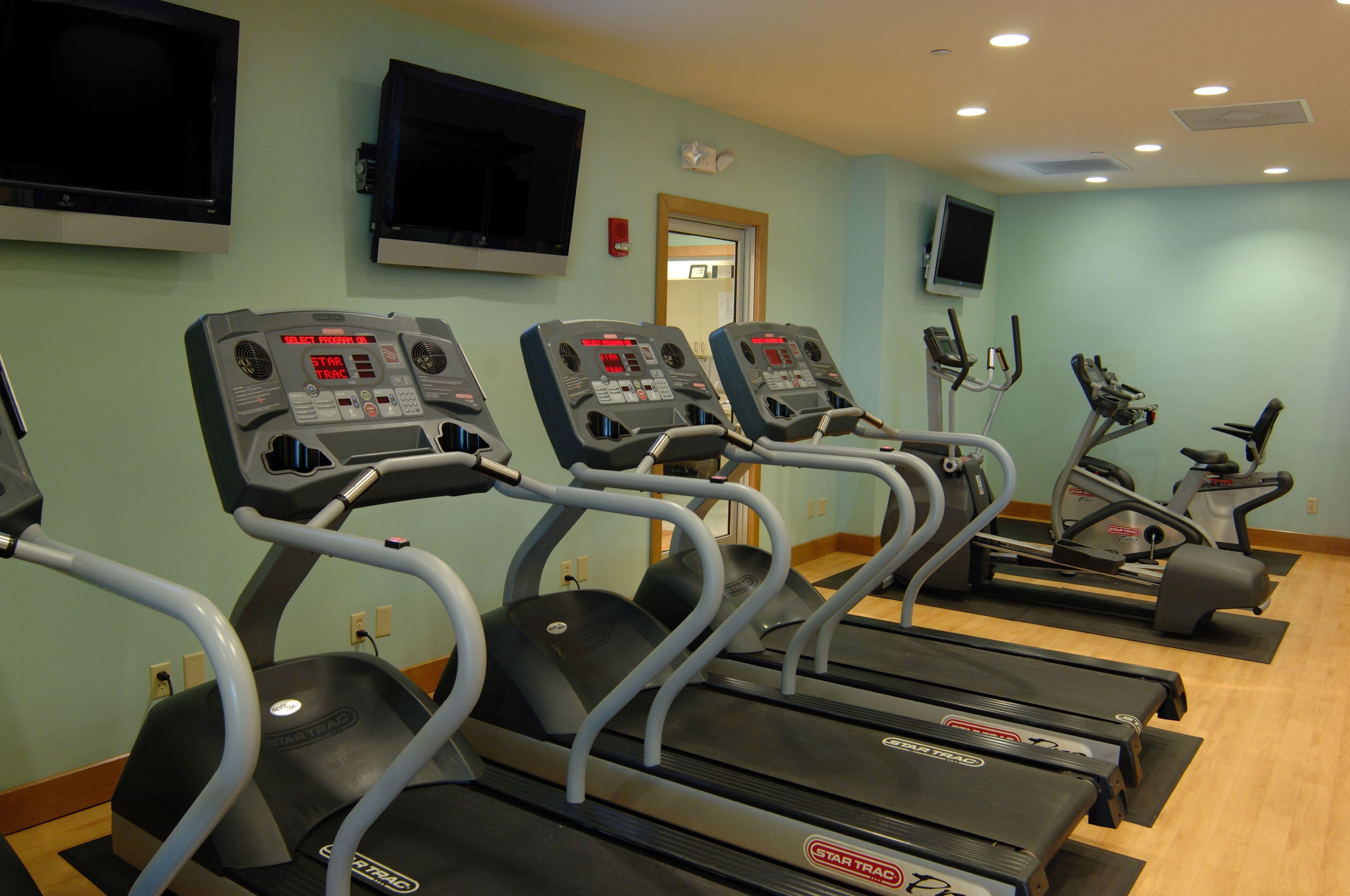 A row of treadmills in a gym with a light green wall, a mounted TV, and a door in the background.
