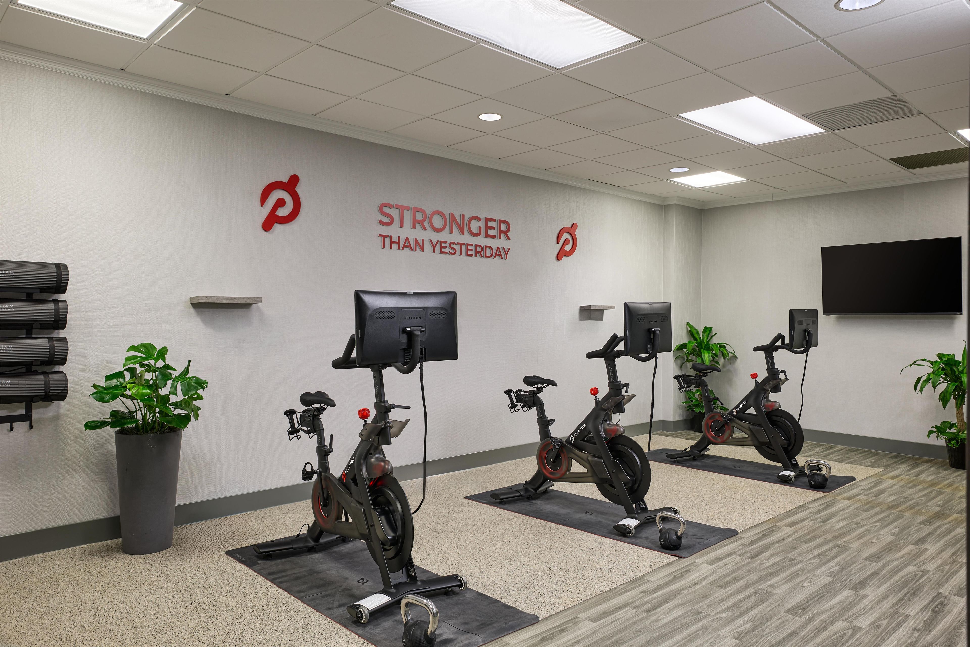 A fitness room with stationary bikes, a motivational wall slogan, a flat-screen TV, and potted plants.