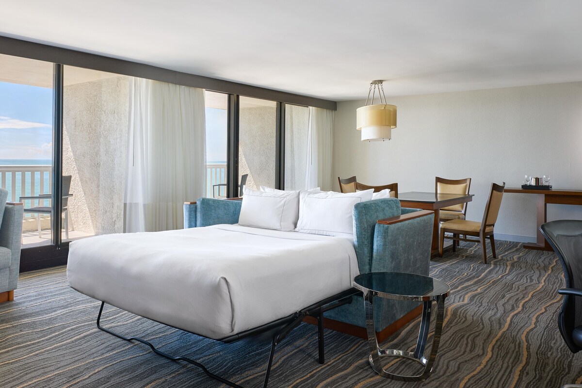 A hotel room with a large bed, a seating area with a table and chairs, a view of the ocean, and a balcony.
