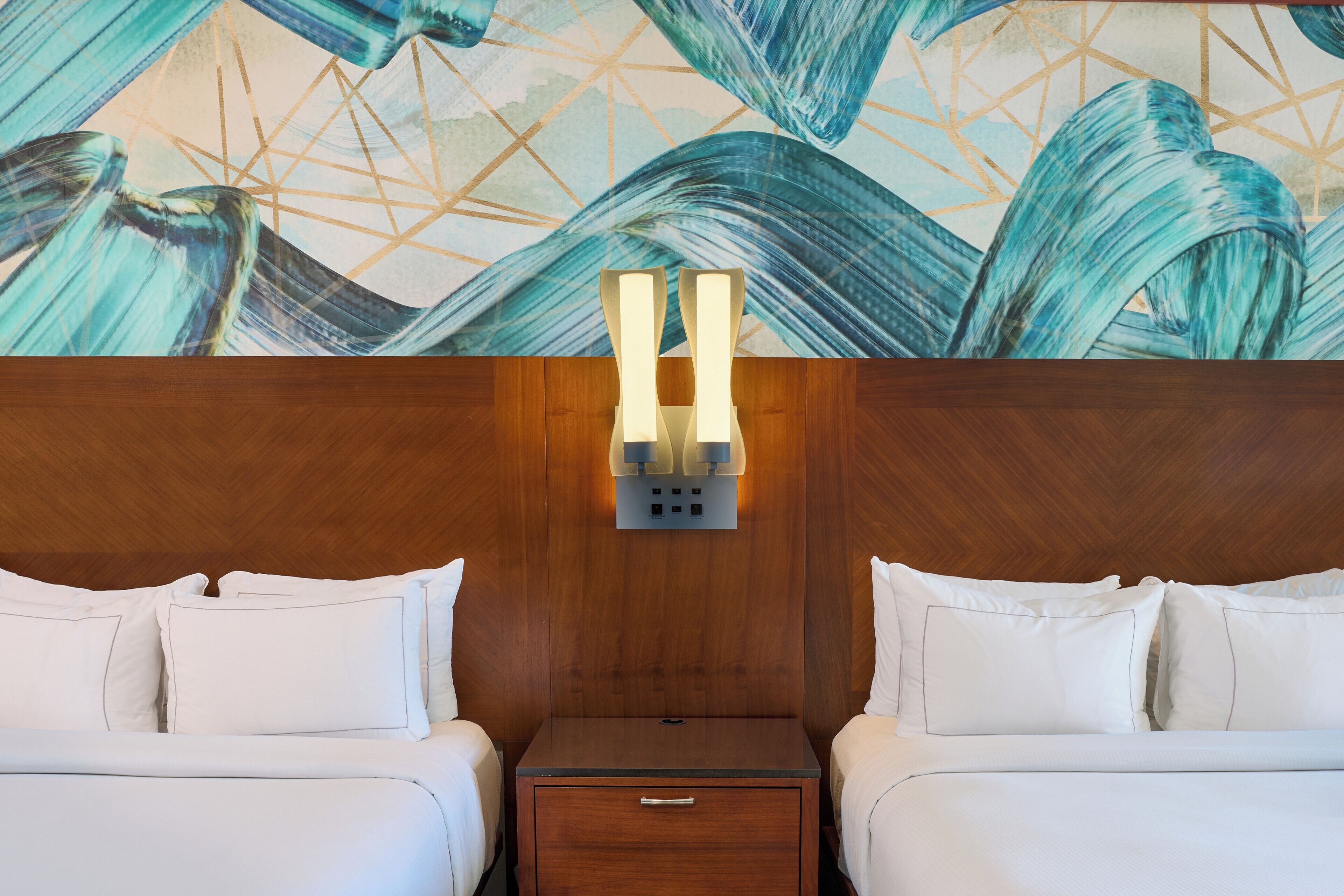 A hotel room with two beds, a wooden headboard, a nightstand with a drawer, and a wall-mounted light fixture.