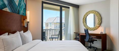 Hotelkamer met een groot bed, een bureau met stoel, een ronde spiegel en uitzicht op gebouwen door een glazen deur.