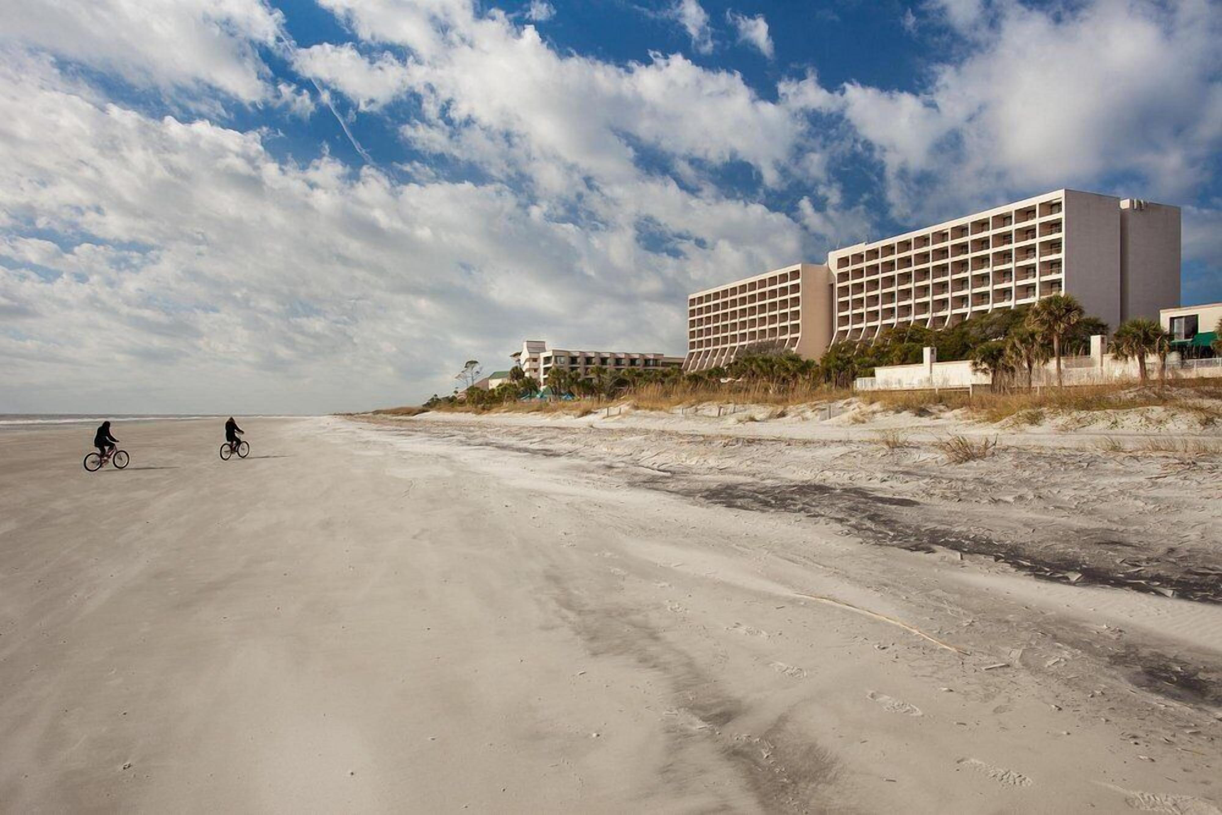 A beachside hotel with a large building, sandy beach, and two cyclists.