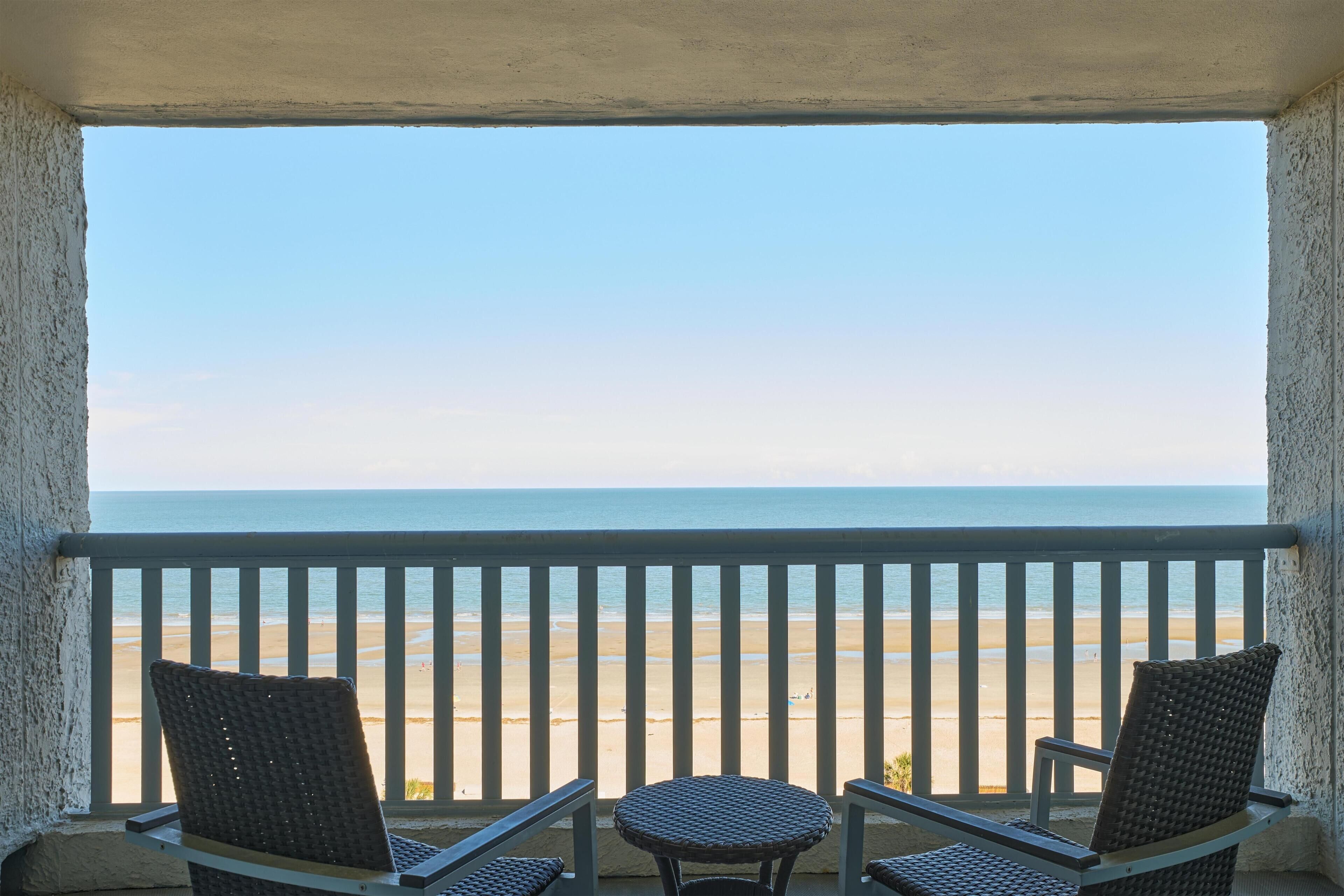A balcony with two wicker chairs and a small table, overlooking a beach.