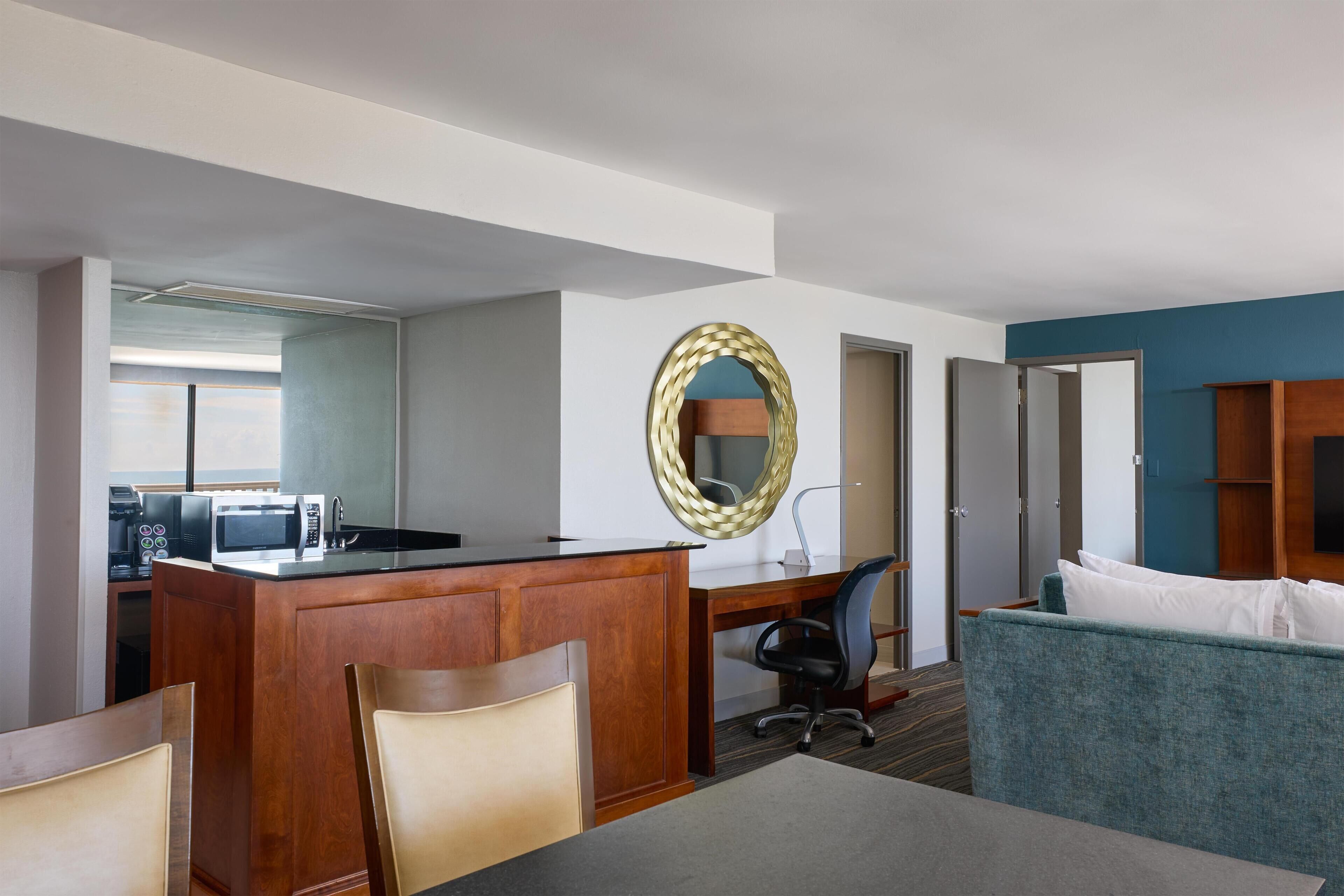 A hotel room with a kitchenette, a desk, a chair, a sofa, and a mirror.