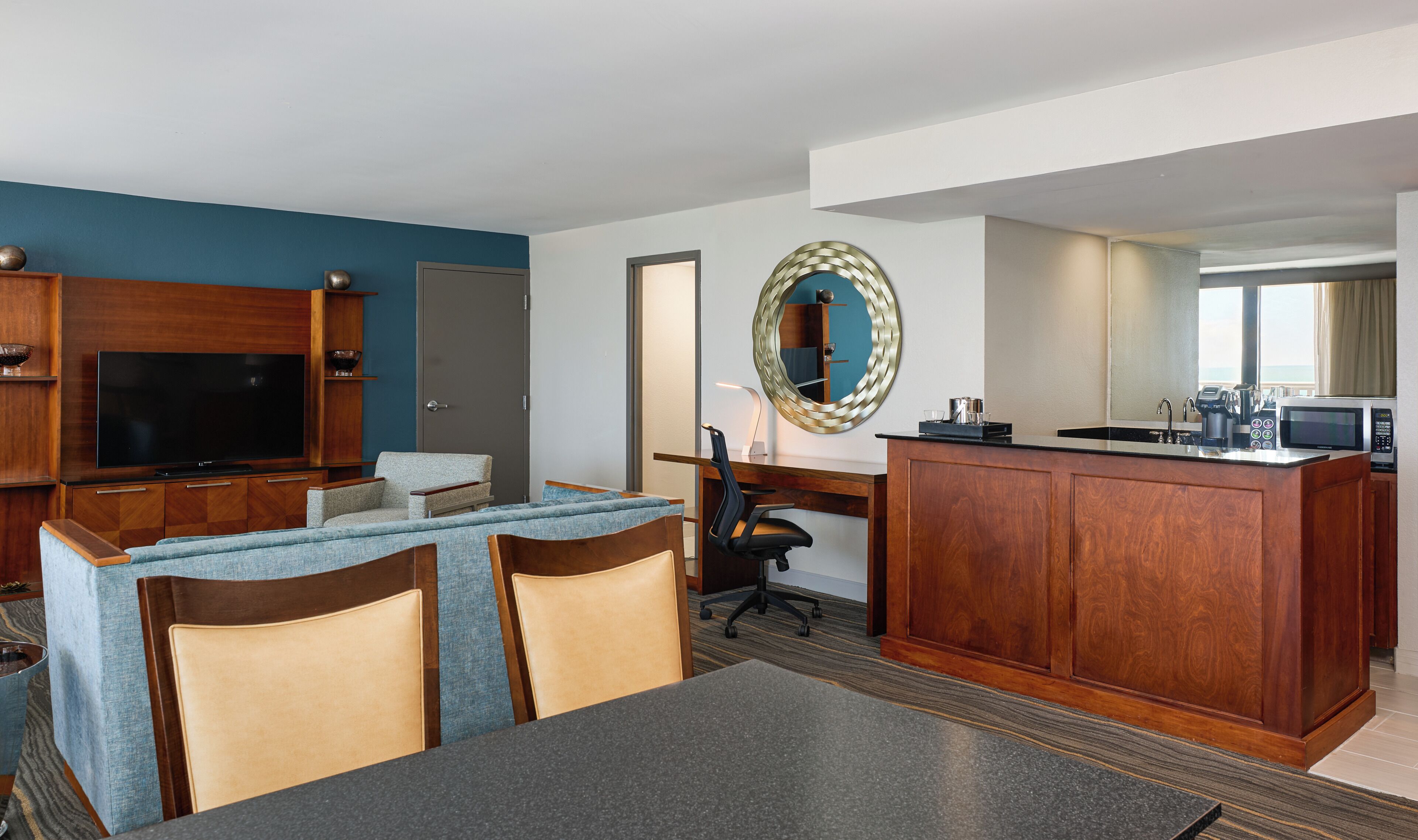 A hotel room with a TV, sofa, dining table, kitchen area, and a large mirror.