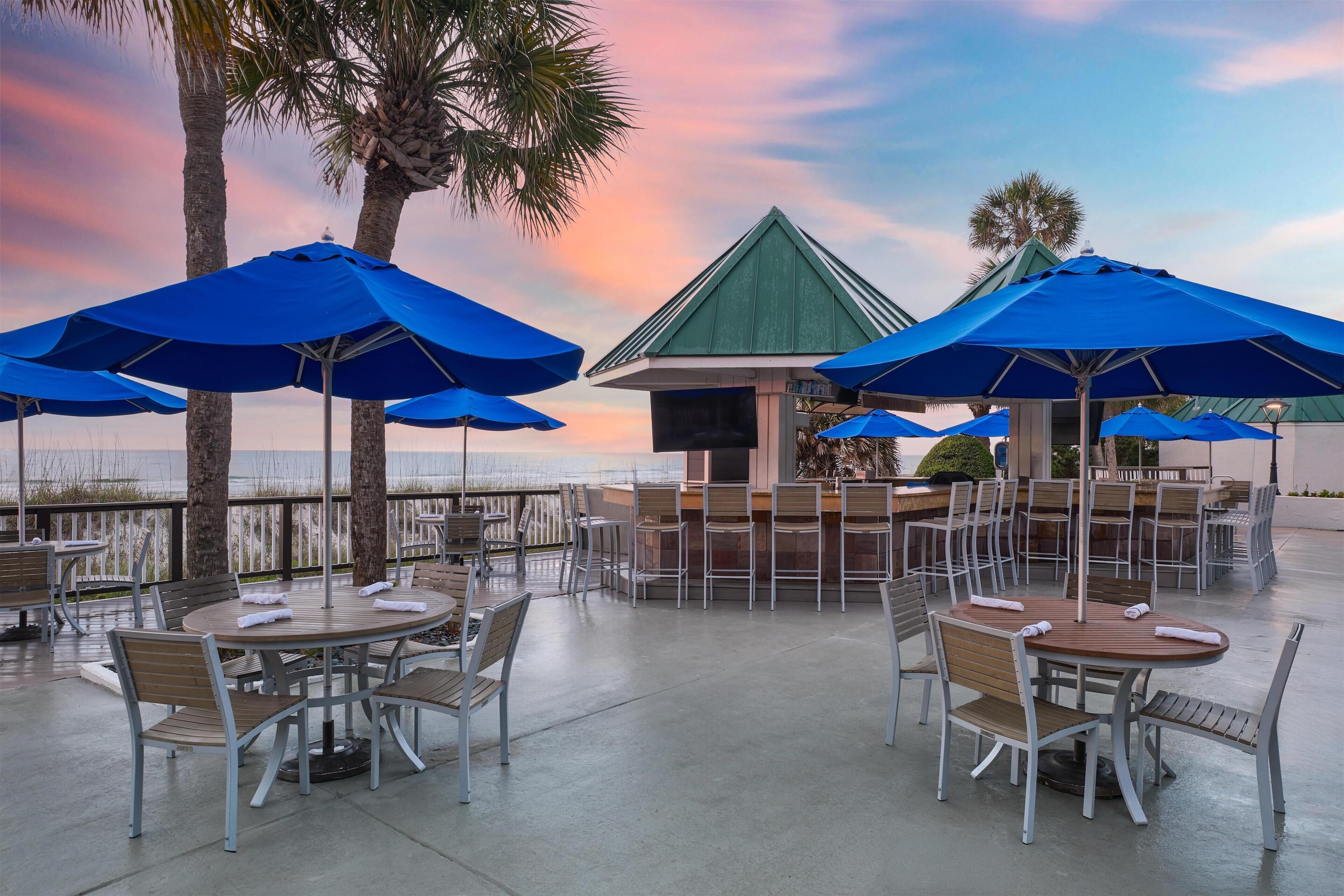Outdoor seating with umbrellas, palm trees, and a view of the water.