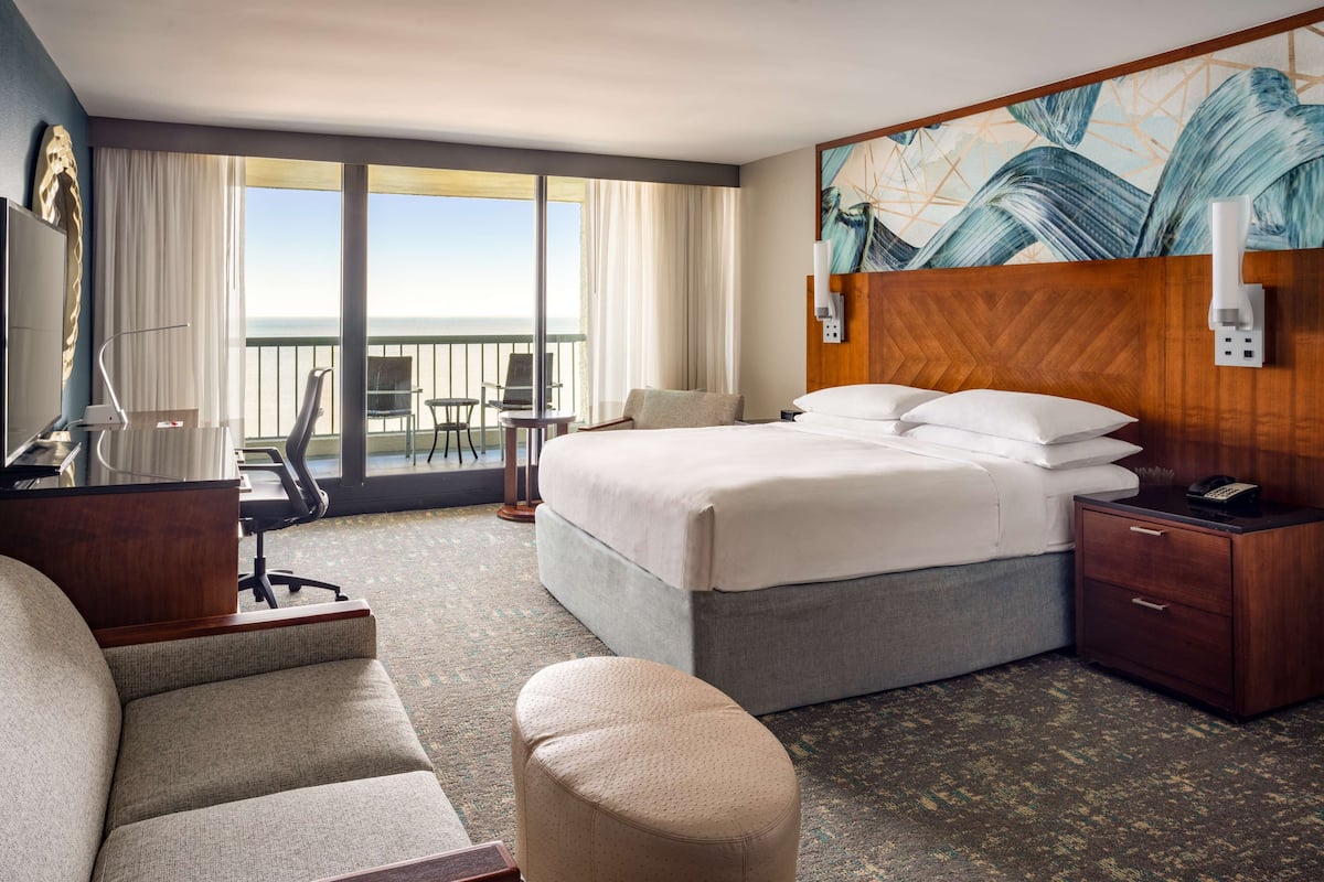 A hotel room with a bed, a desk, a chair, a small table, a sofa, and a balcony with a view of the ocean.