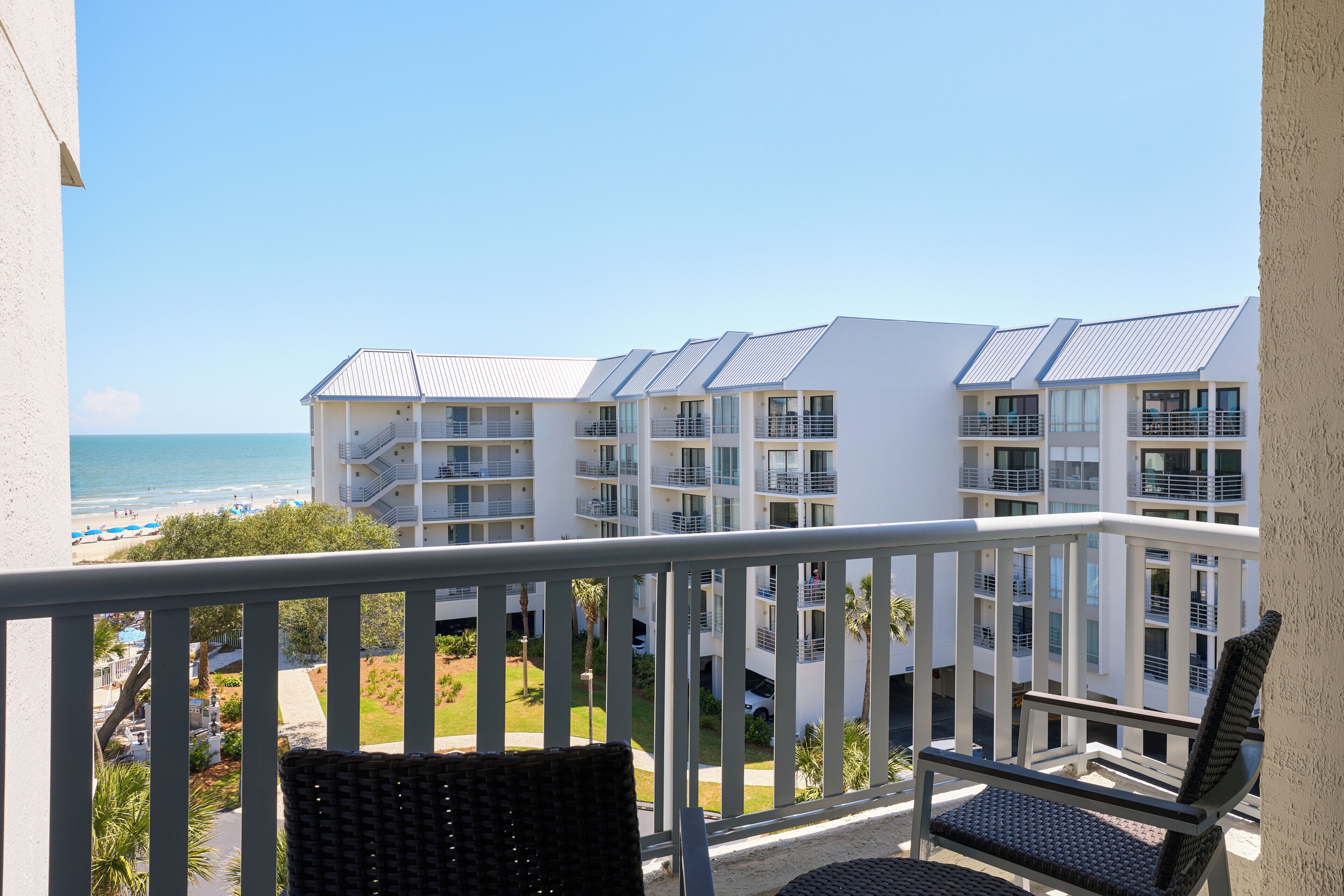 Un balcon agrémenté de fauteuils en osier, offrant une vue sur une plage et un bâtiment d’hôtel doté de nombreux balcons.