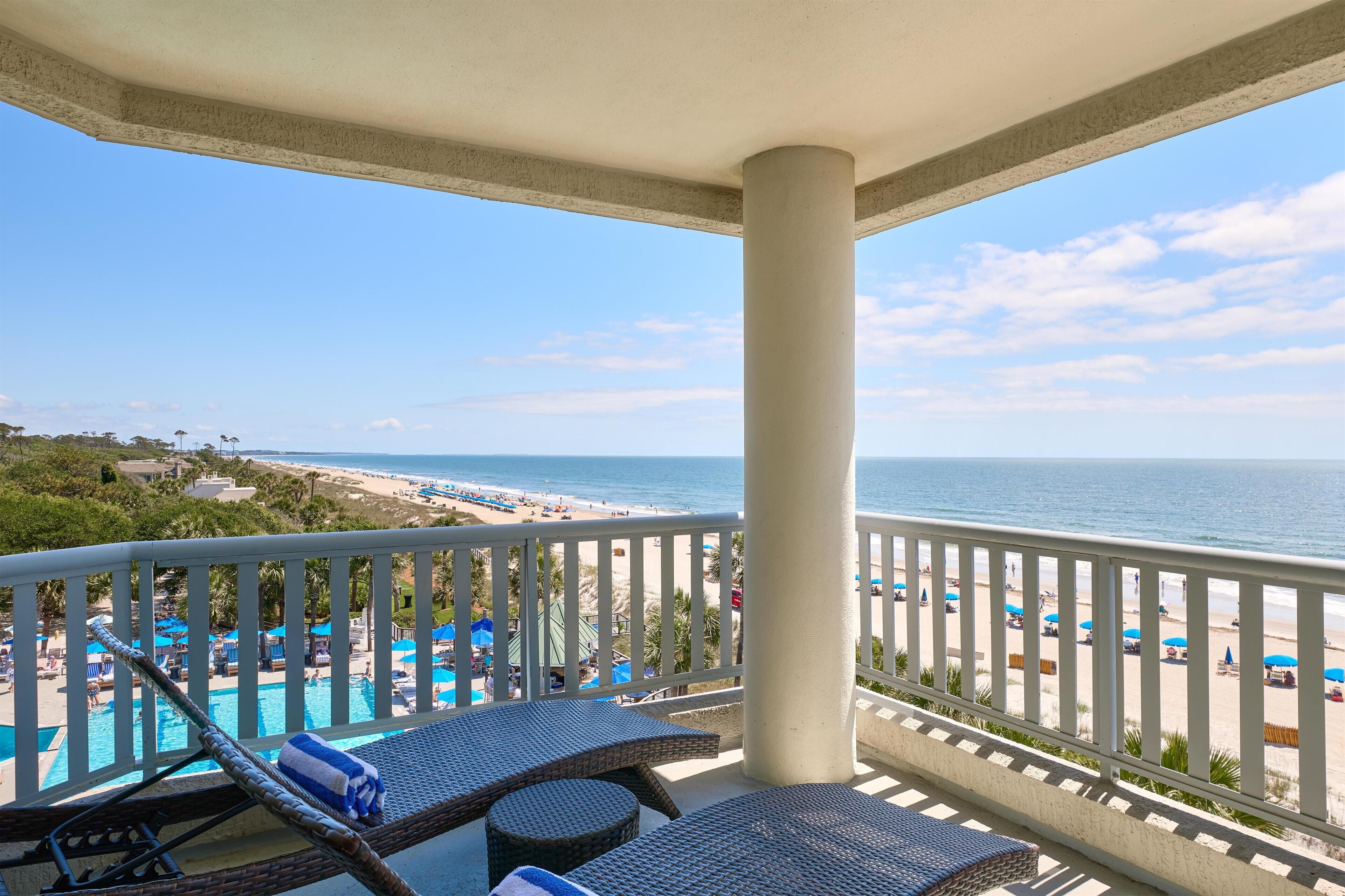 A balcony with wicker lounge chairs, a pool, and a view of the beach and ocean.