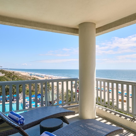 A balcony with wicker lounge chairs, a pool, and a view of the beach and ocean.