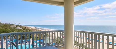 A balcony with wicker lounge chairs, a pool, and a view of the beach and ocean.