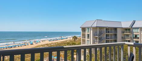 A balcony with a view of a beach and ocean, a pool, and a building with multiple balconies.