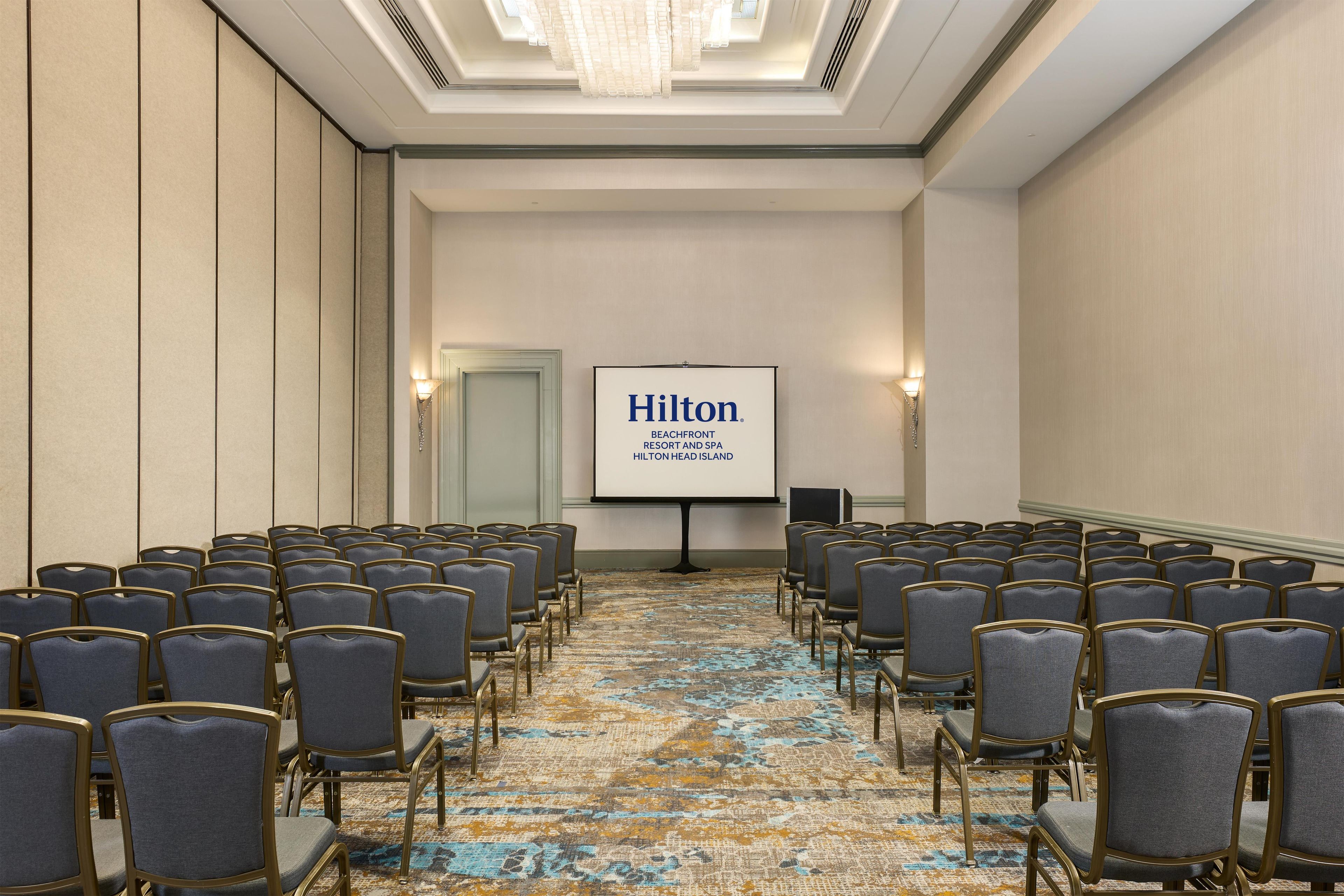 Un salón de conferencias con filas de sillas, una pantalla de proyector mostrando 'Hilton Beachfront Resort and Spa' y un piso alfombrado.