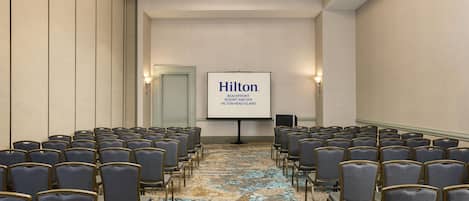 Un salón de conferencias con filas de sillas, una pantalla de proyector mostrando 'Hilton Beachfront Resort and Spa' y un piso alfombrado.