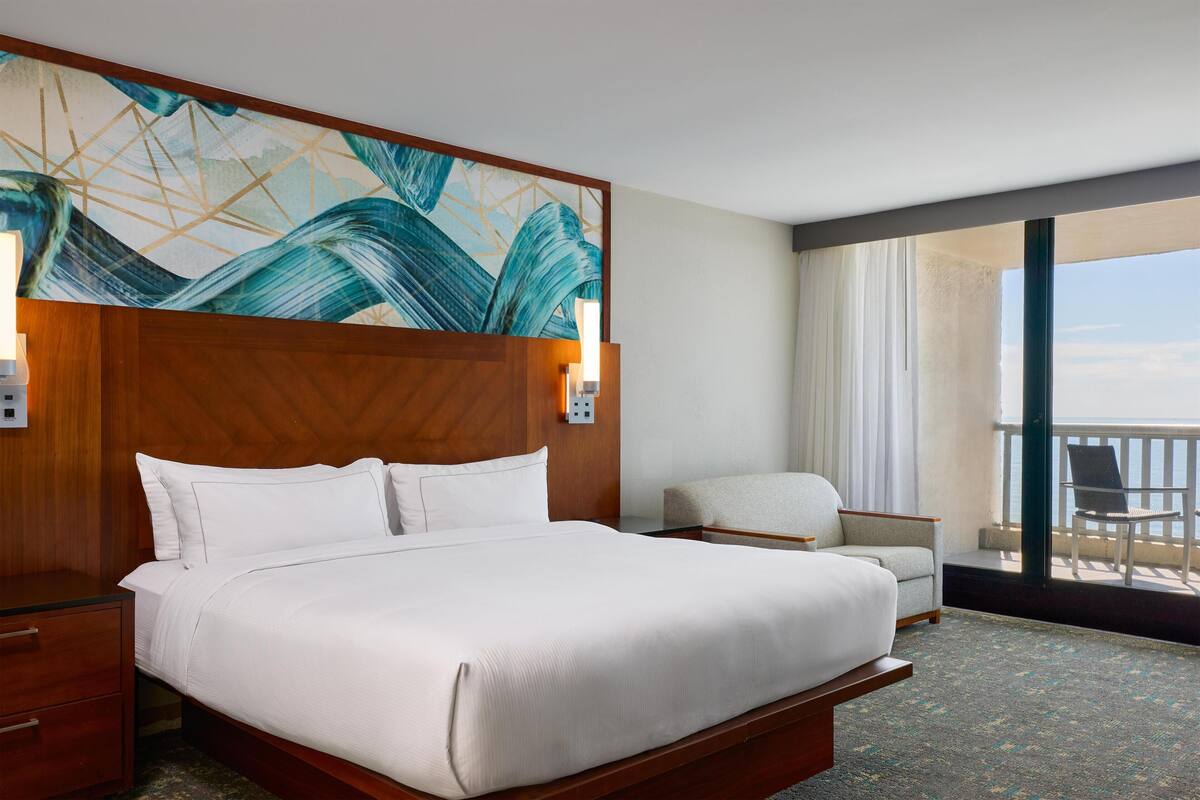 A hotel room with a large bed, a wooden headboard, a painting, a chair, and a balcony with a view.