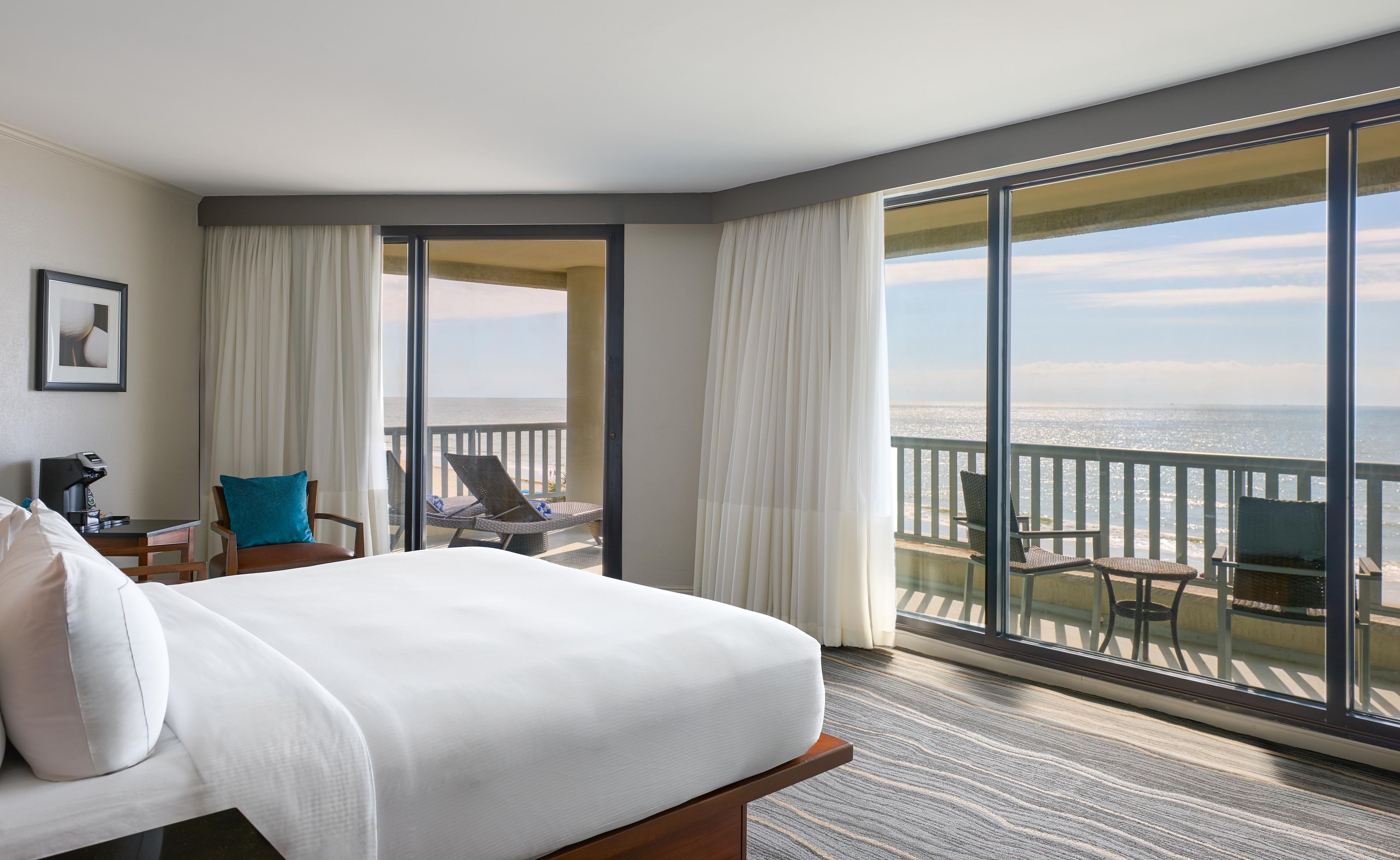 A hotel room with a large bed, a desk with a coffee machine, a chair, and a balcony with a view of the ocean.