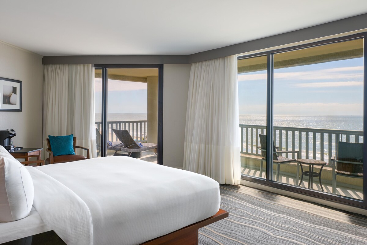 A hotel room with a large bed, a desk with a coffee machine, a chair, and a balcony with a view of the ocean.