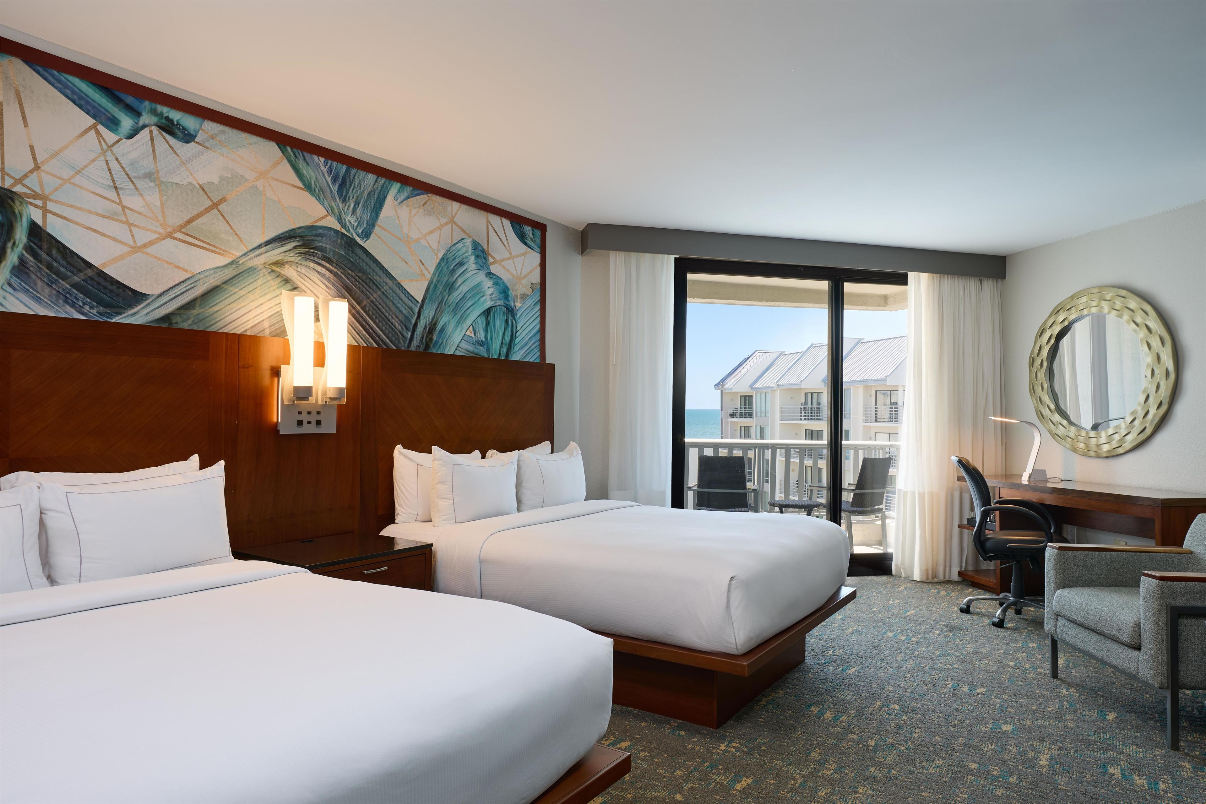 A hotel room with two beds, a desk, a chair, a large window with a view, and a decorative wall piece.