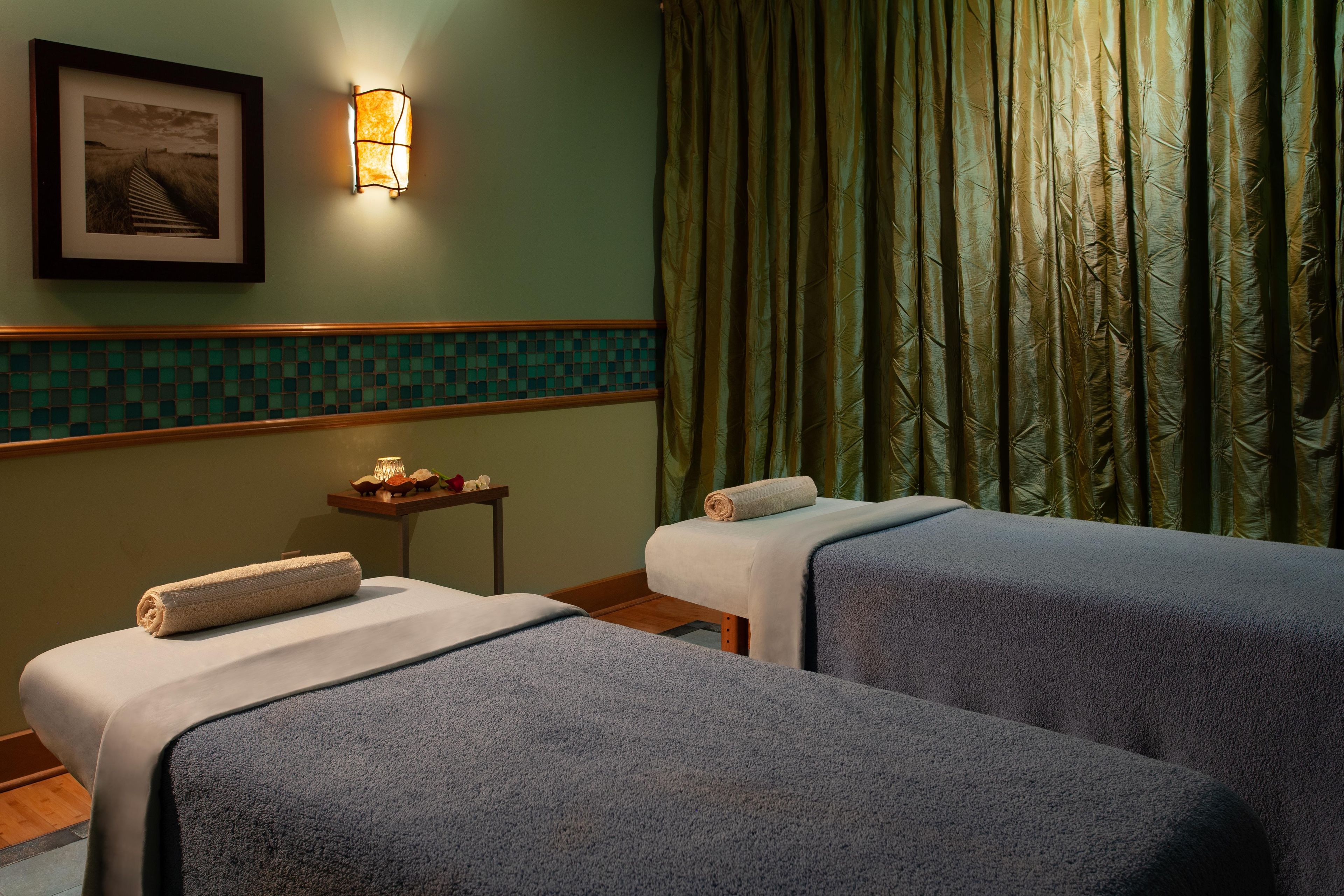 A room with two beds, a small table with a lamp, a picture on the wall, and green curtains.