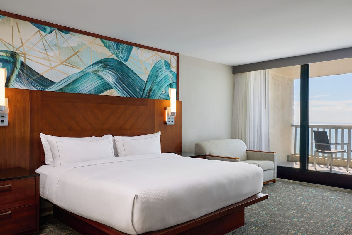A hotel room with a large bed, a nightstand, a chair, a view of the ocean, and a painting on the wall.