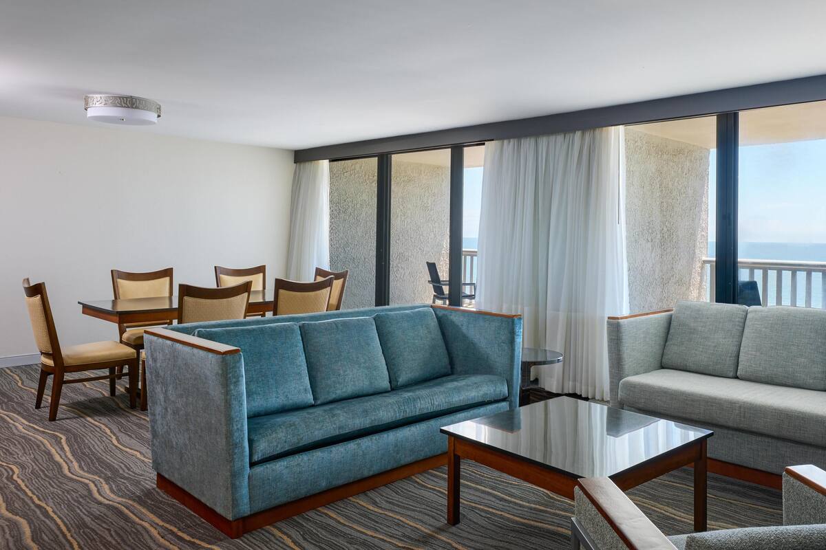 A hotel room with a seating area, a dining table, and large windows with a view.