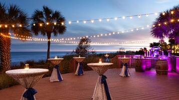 A deck with tables and chairs, overlooking a beach with palm trees and string lights.