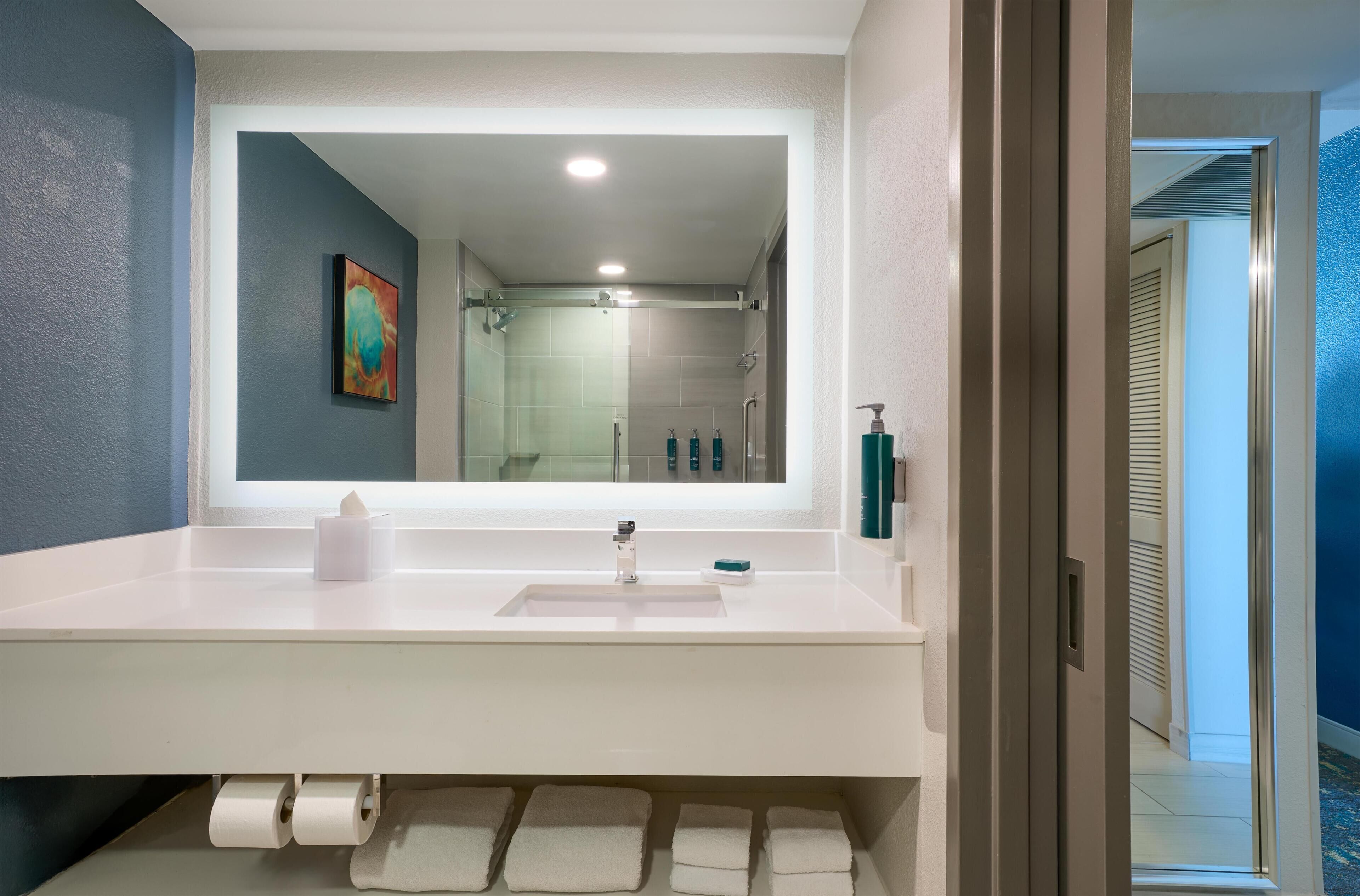 Un bagno moderno con uno specchio grande, un lavandino bianco e una zona doccia visibile attraverso lo schermo di vetro.